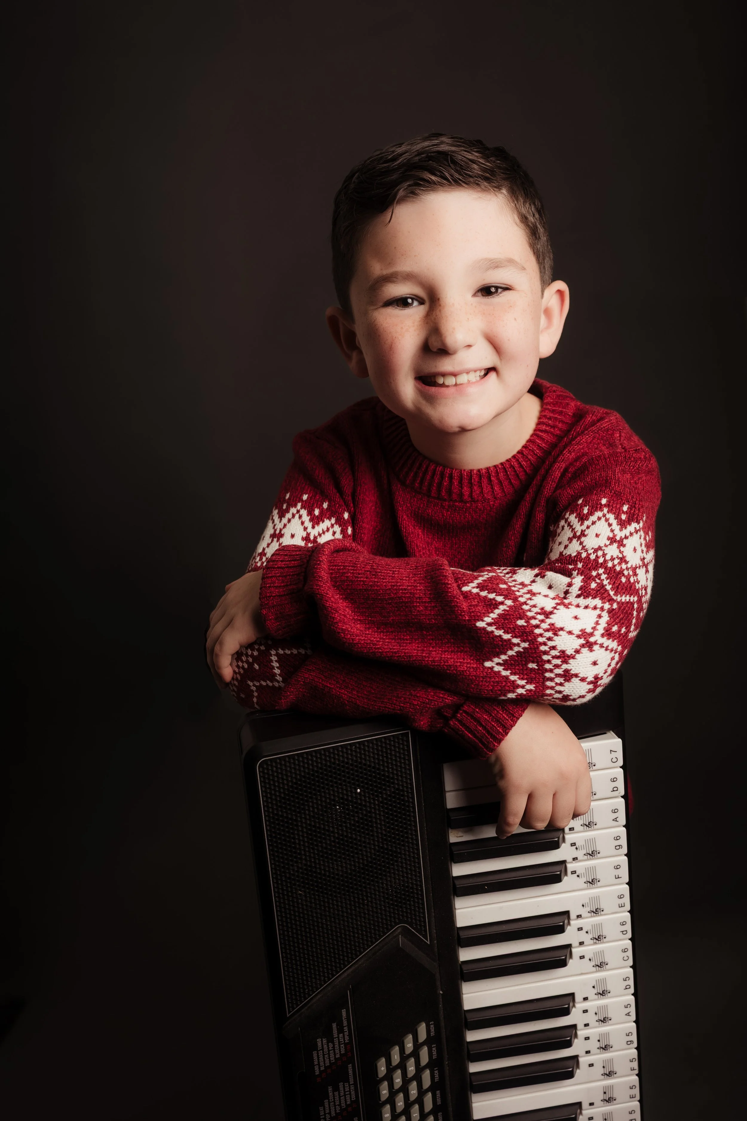 A young boy with short brown hair, smiling, wearing a red holiday sweater with white snowflake patterns, resting his arms on a black and white electronic keyboard, against a dark background.