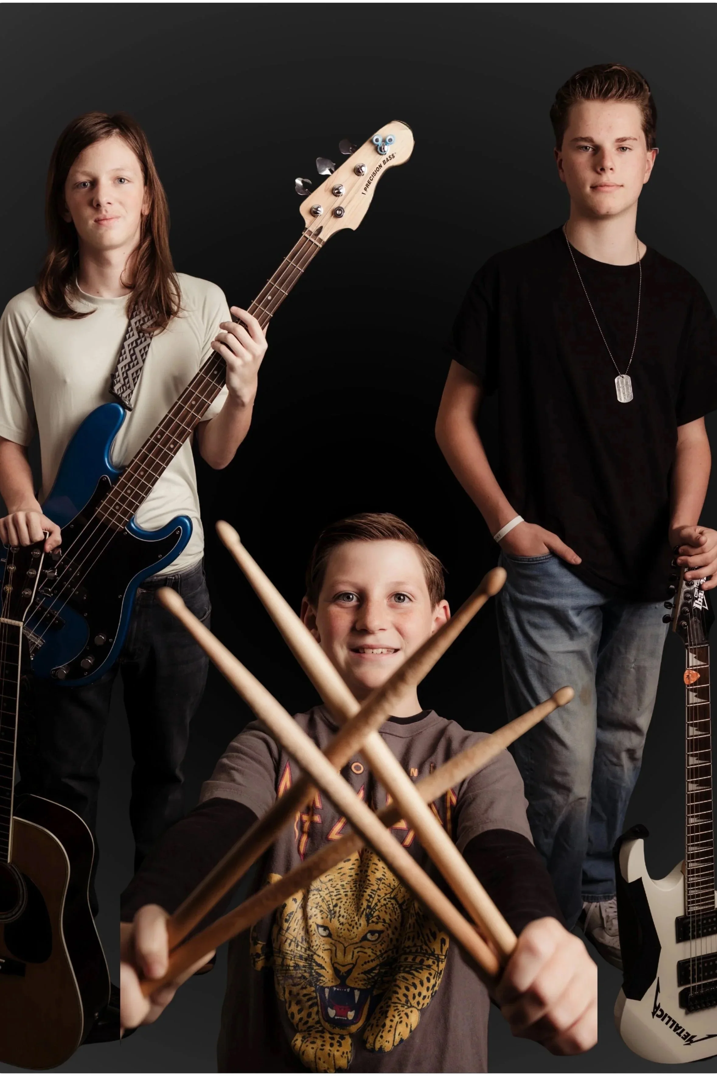 Three young males with musical instruments on a dark background, one with an electric bass, one with an electric guitar, and the third holding drumsticks.