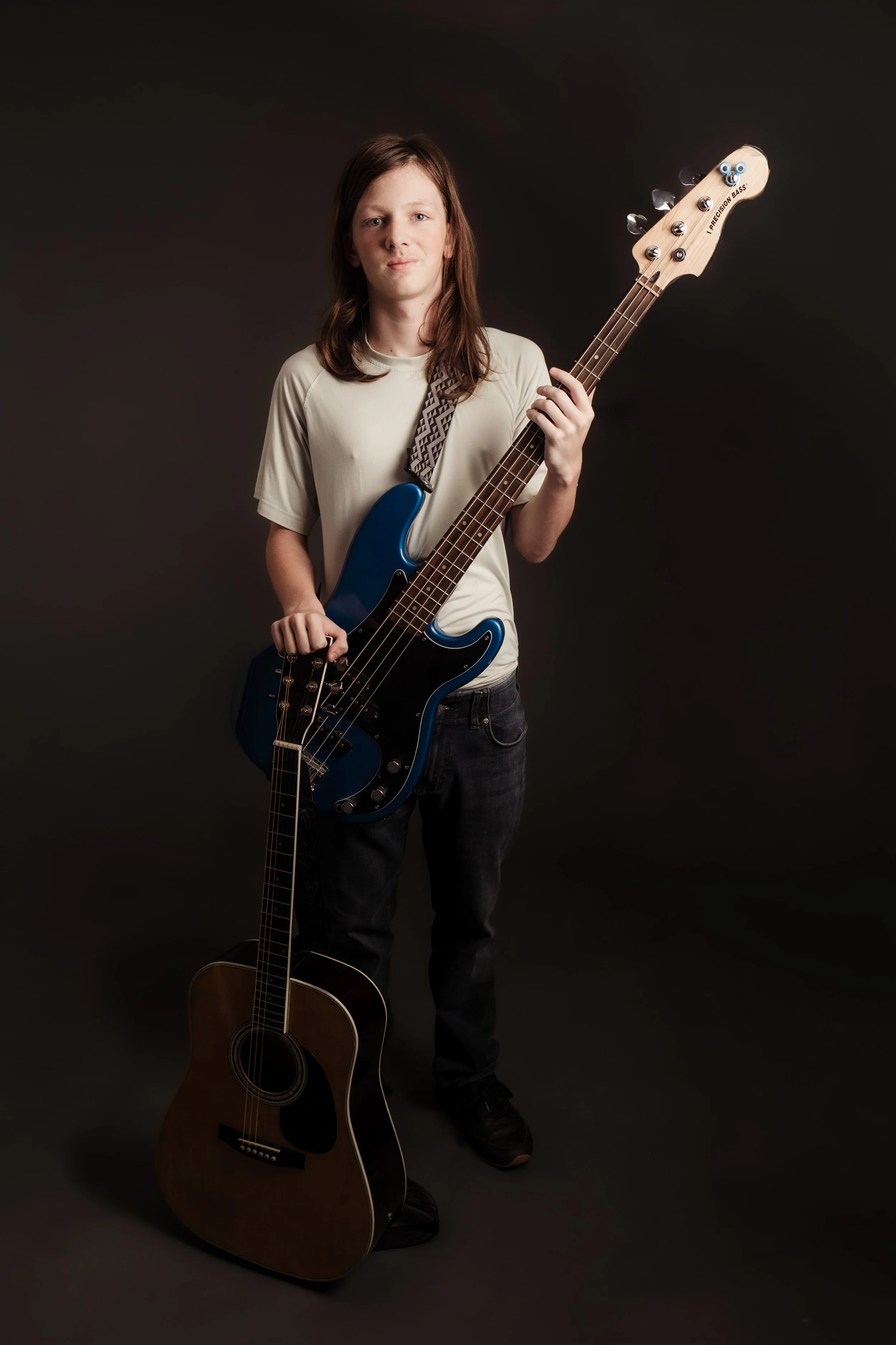 A young person standing against a dark background, holding a blue electric bass guitar, with an acoustic guitar resting on the floor.