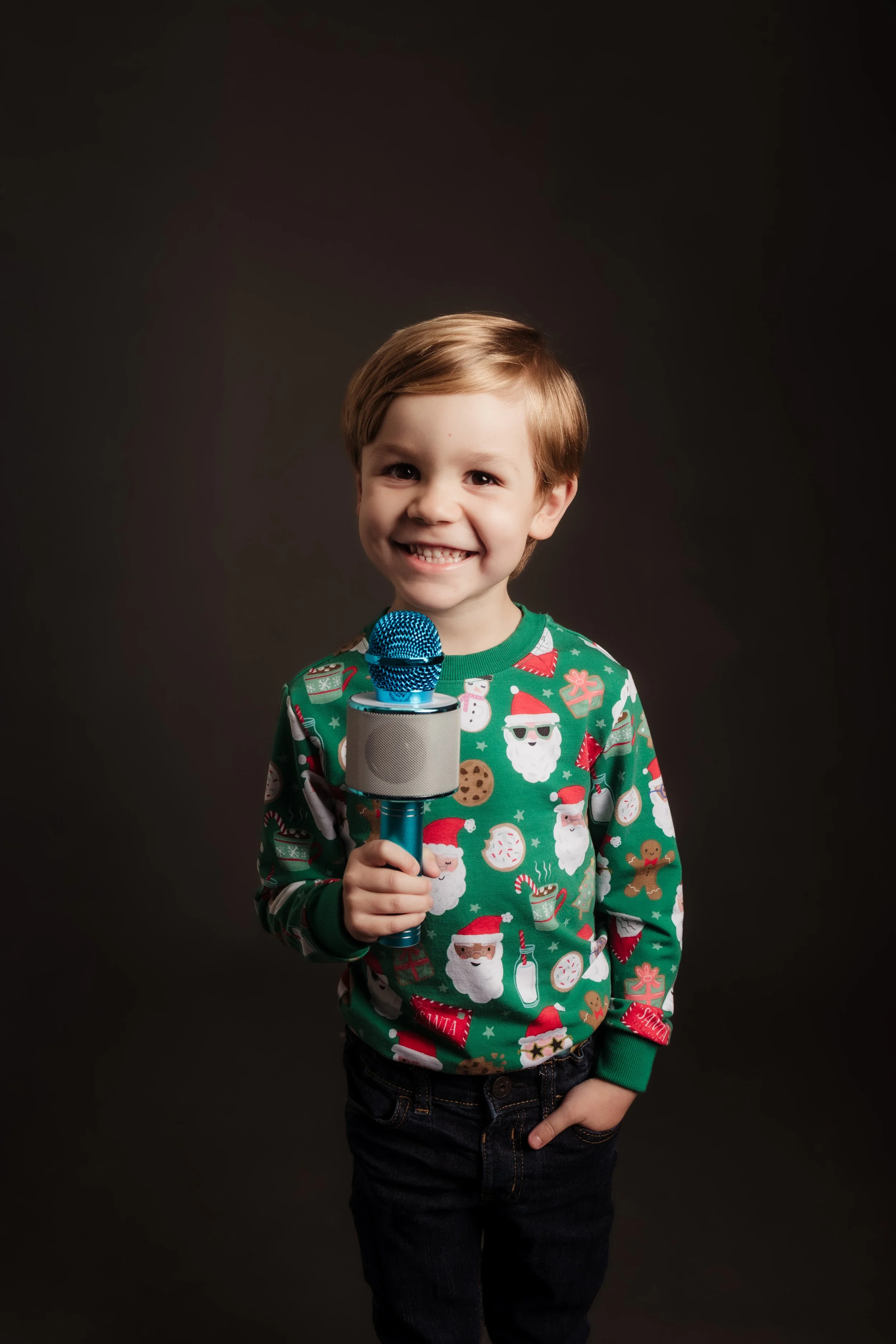 A young boy with light brown hair smiling and holding a microphone, wearing a Christmas sweater featuring Santa Claus, cookies, and holiday-themed decorations, against a dark backdrop.