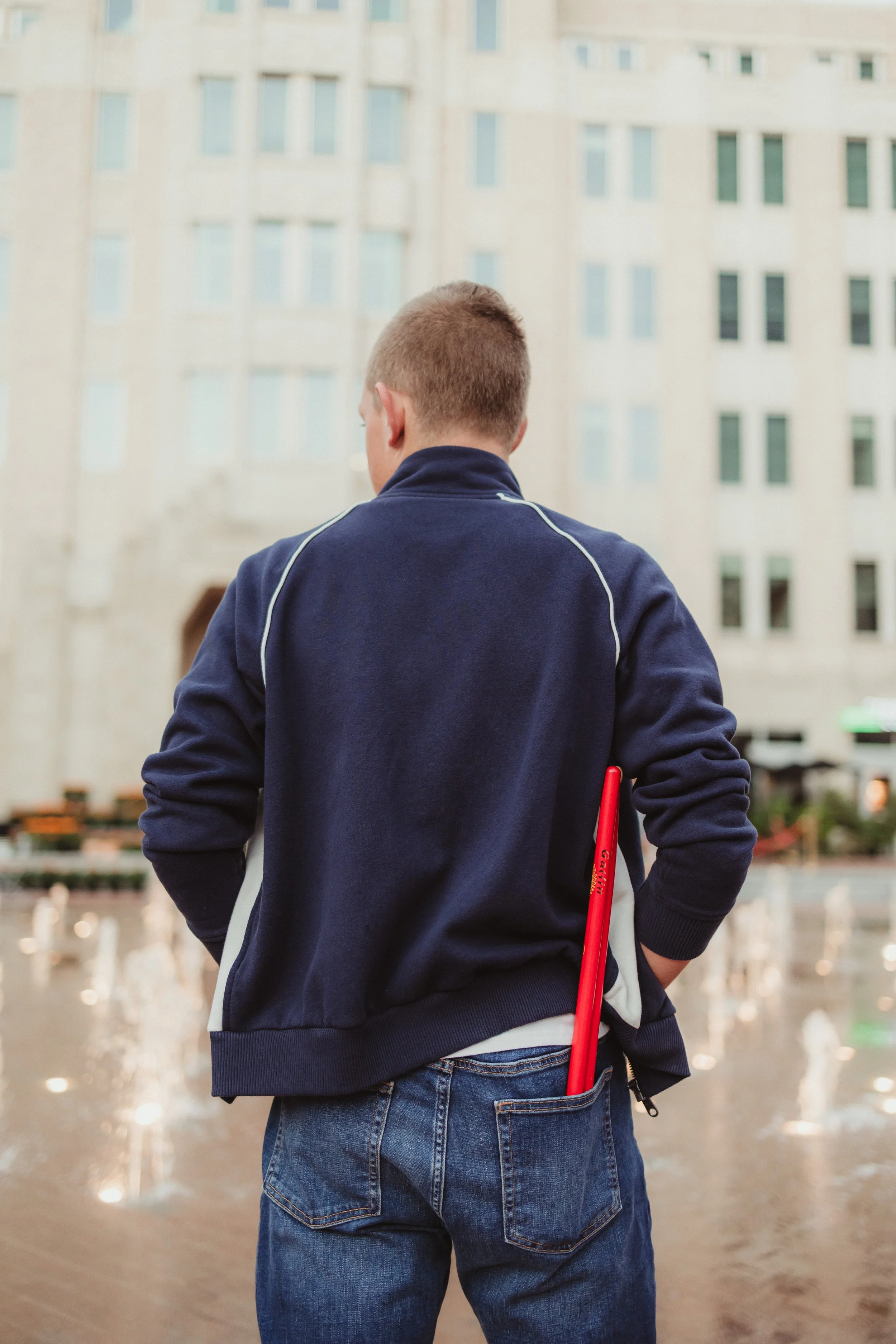 Back of a young man in a navy jacket and jeans standing in front of a building and fountain, holding a red object.