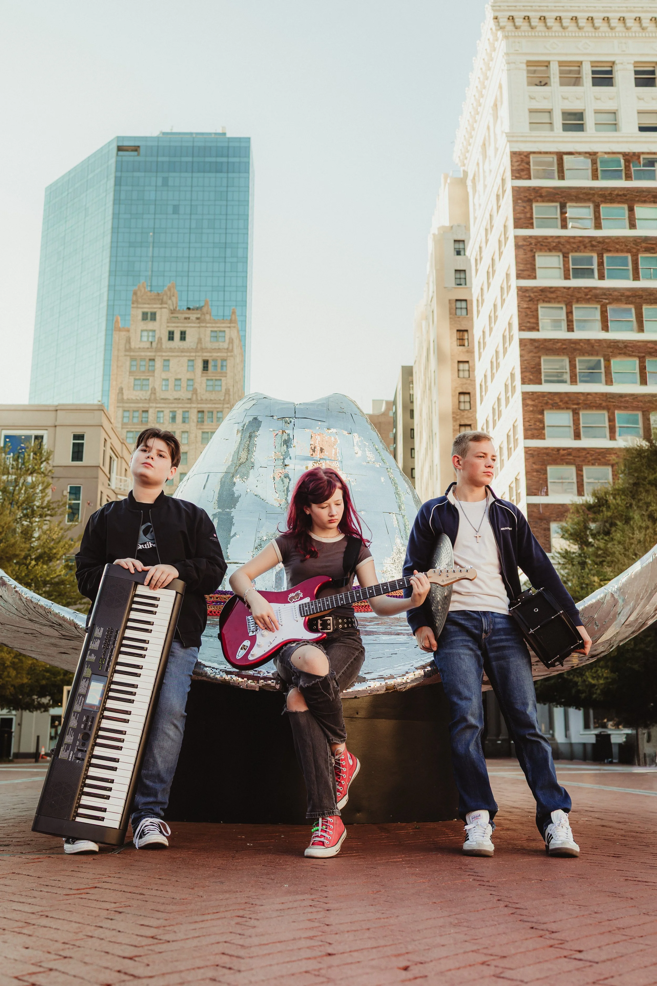 Three young people with musical instruments standing in front of a public art installation in an urban park, with skyscrapers in the background.