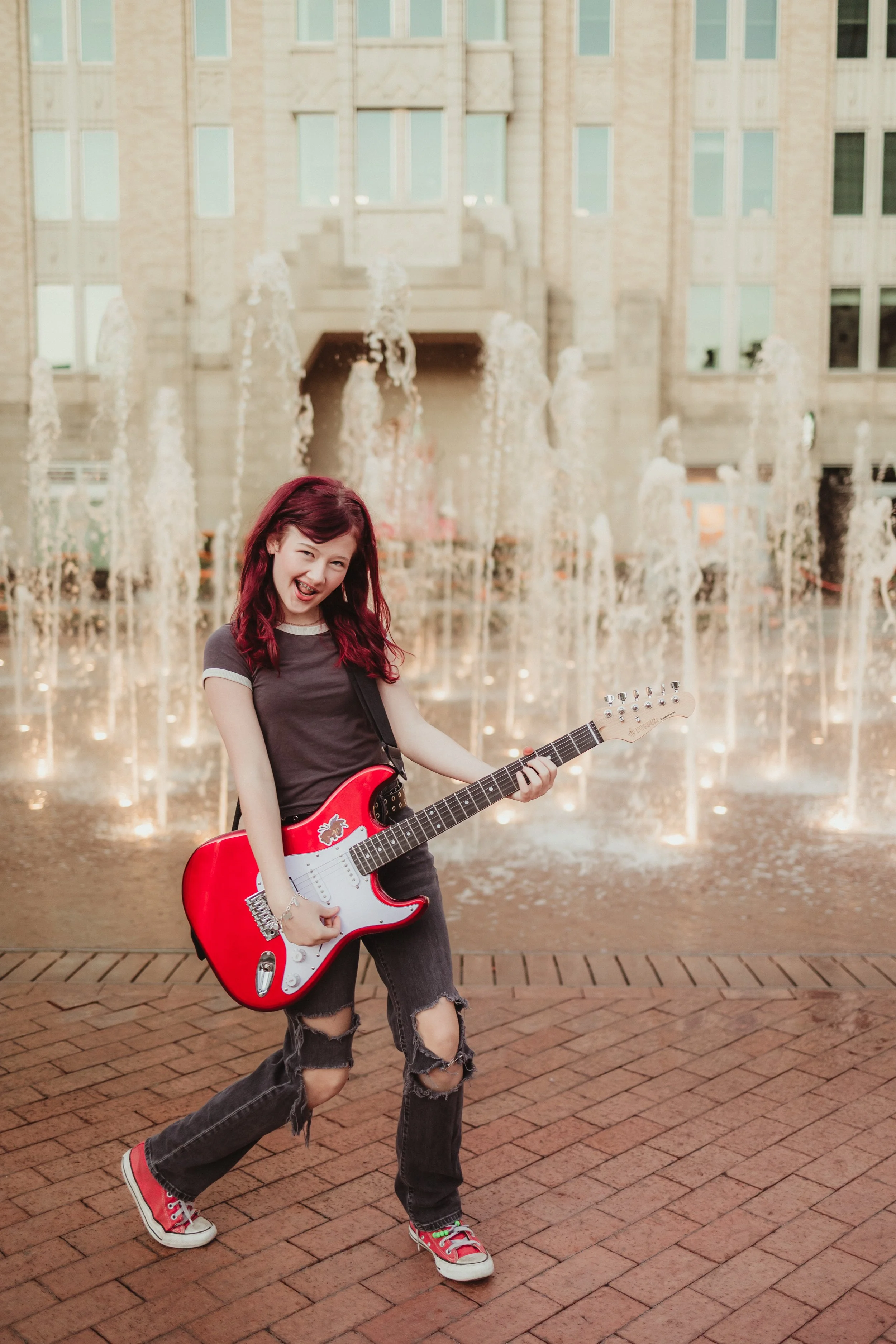 Young woman with red hair playing a red electric guitar by a fountain in front of a large building.