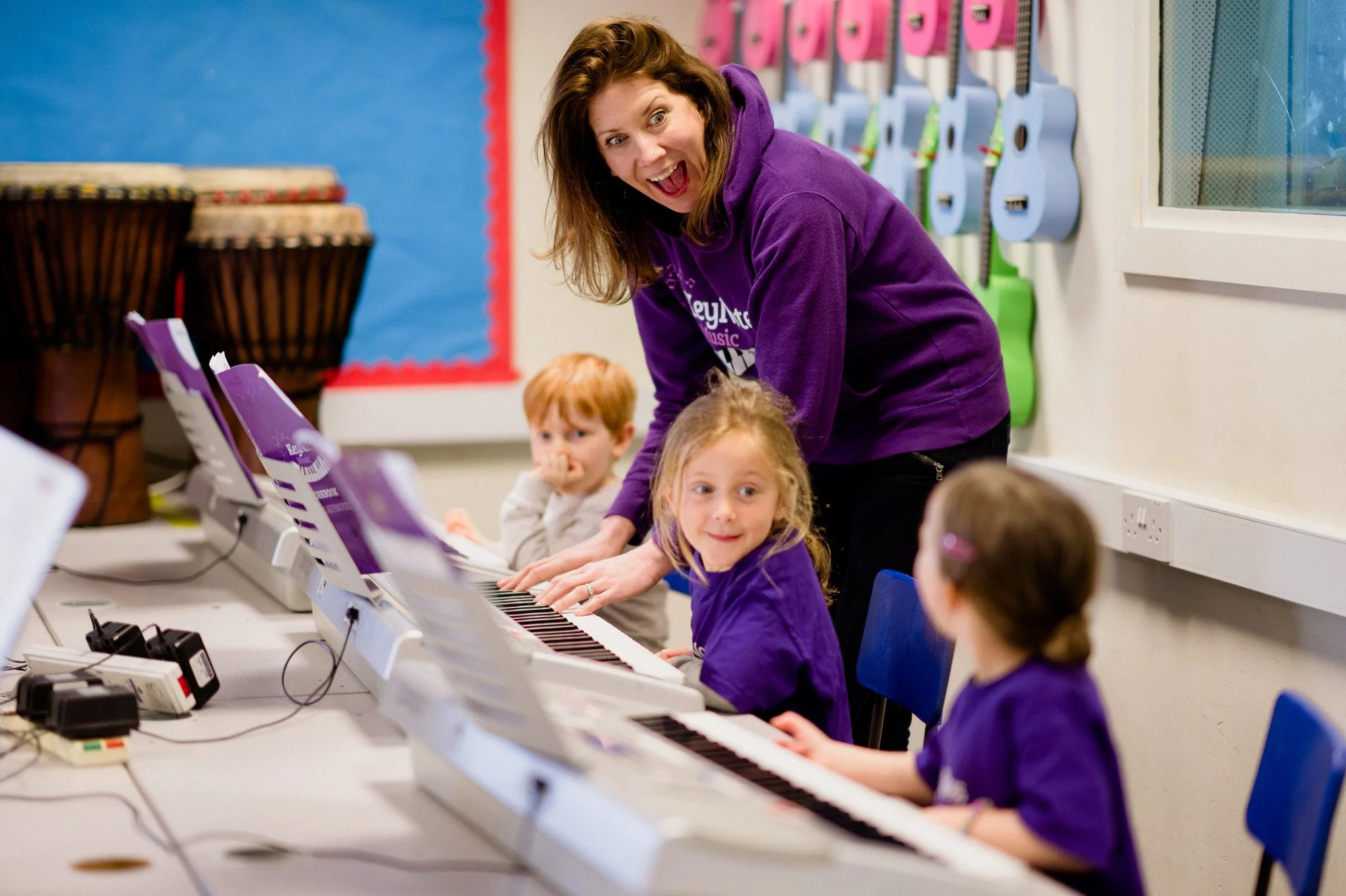 Music teacher guiding young children in a music classroom, with children playing keyboards and guitars on the wall.