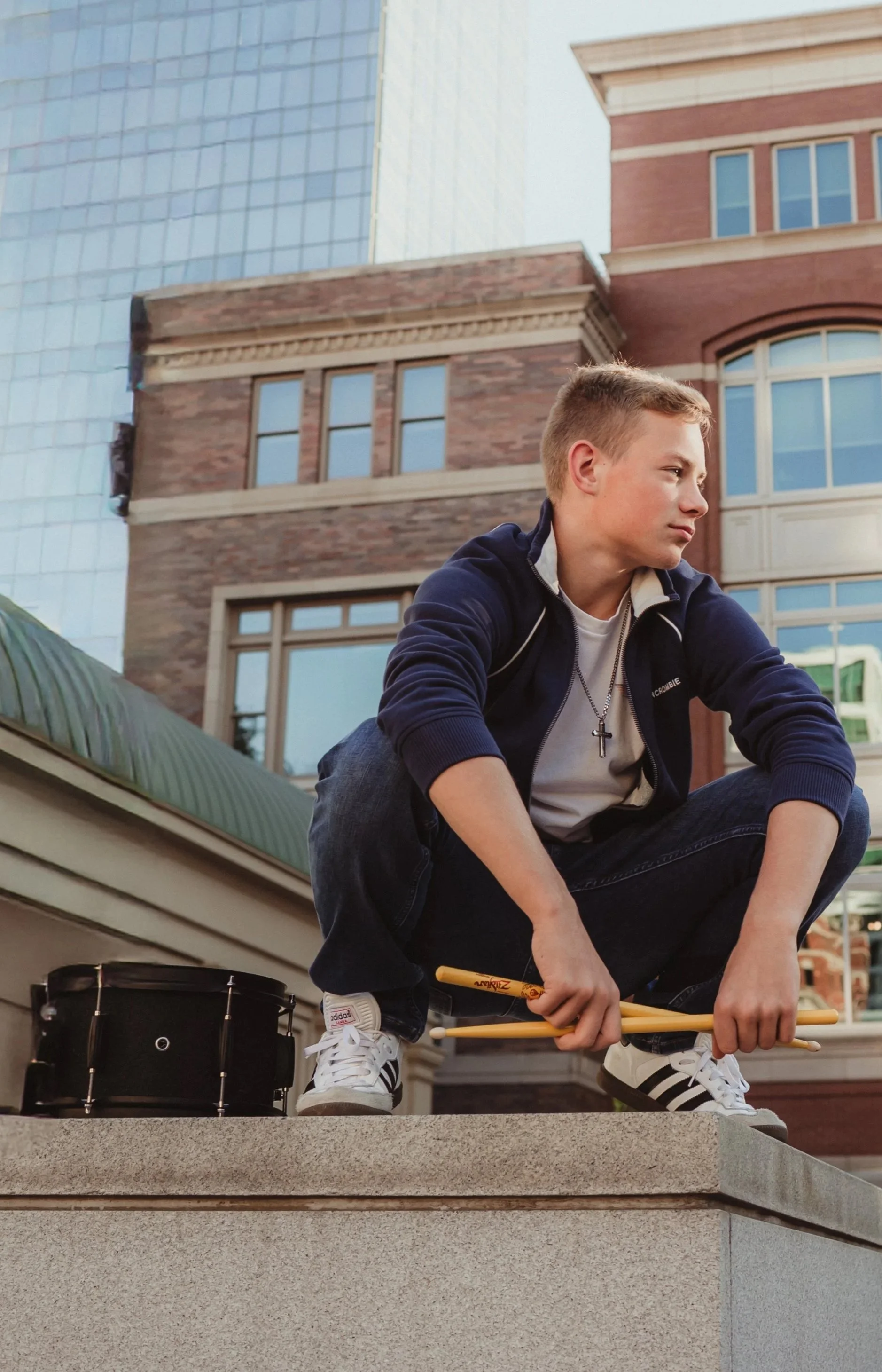 Young man crouching on a rooftop with a microphone and a drum, looking contemplative, with city buildings in the background.