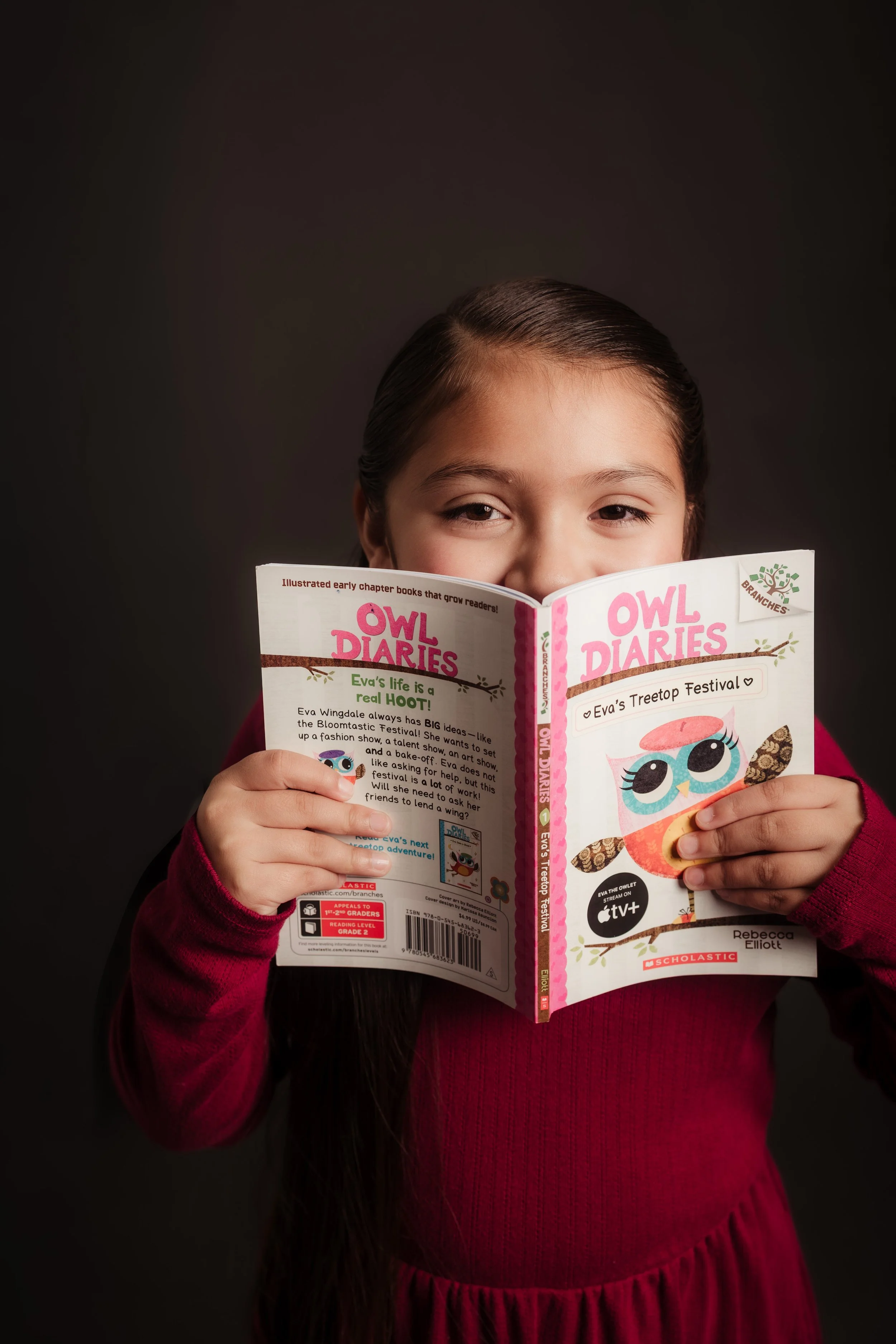 A young girl with long dark hair and a red sweater reads a colorful book titled 'Owl Diaries' with a cute owl illustration on the cover. The background is plain and dark.