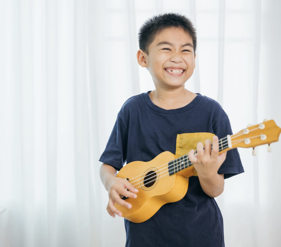 A smiling boy in a navy T-shirt playing a small yellow ukulele in a bright room.