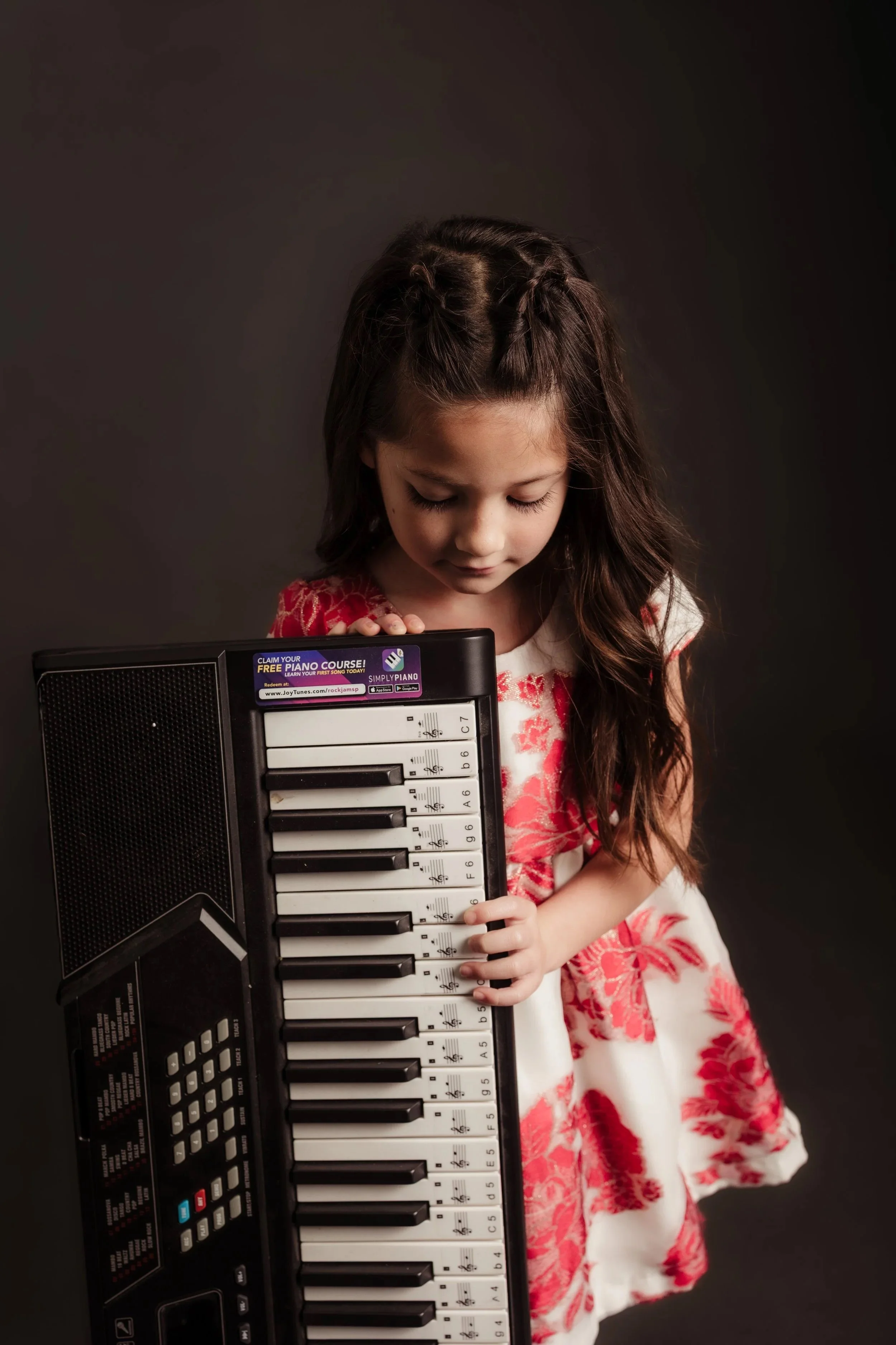 A young girl with long brown hair standing next to a small electronic keyboard, looking down at it.