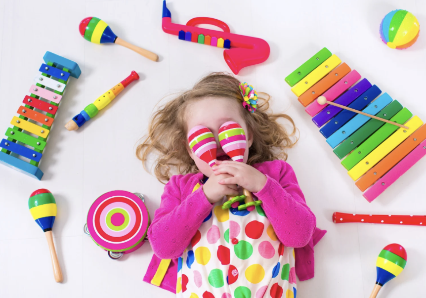 Child lying on a white surface surrounded by colorful musical toys, holding a pair of striped maracas over her face, wearing a polka dot dress and pink cardigan.