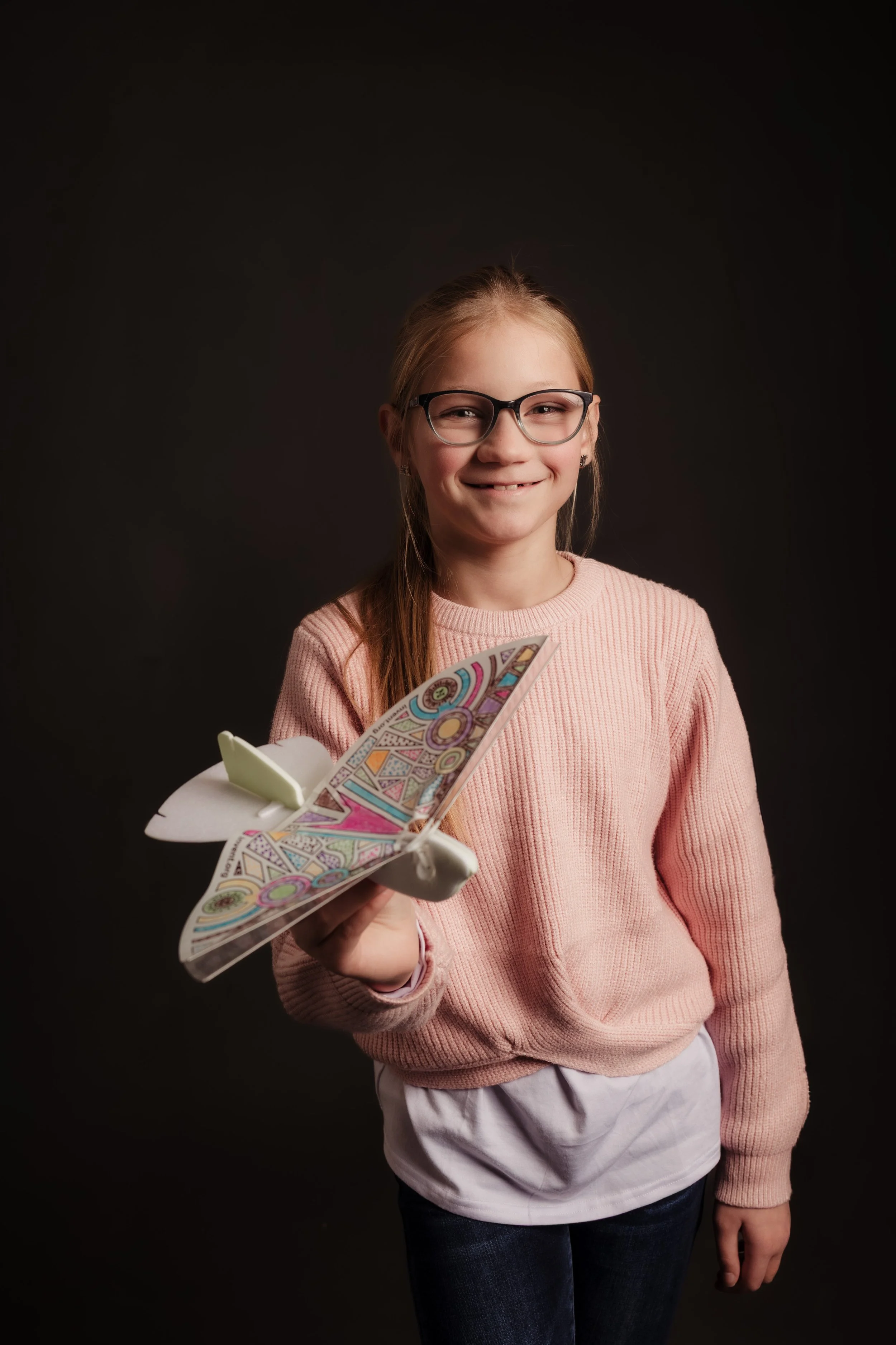 A young girl with glasses wearing a pink sweater, holding a colorful paper butterfly craft, smiling at the camera against a dark background.