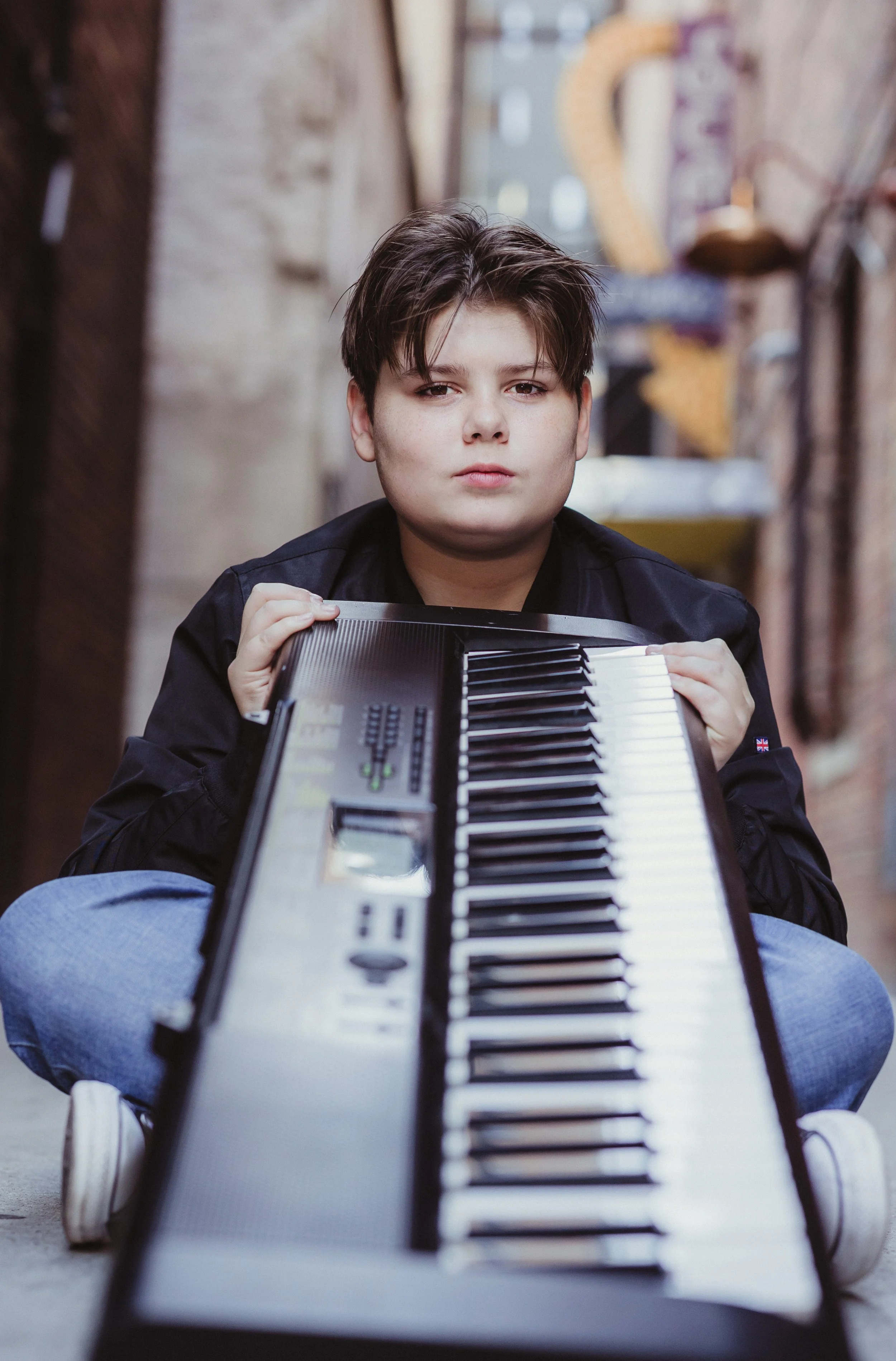 A young person holds a keyboard piano, sitting outdoors between brick and wooden walls, with an urban alley background.