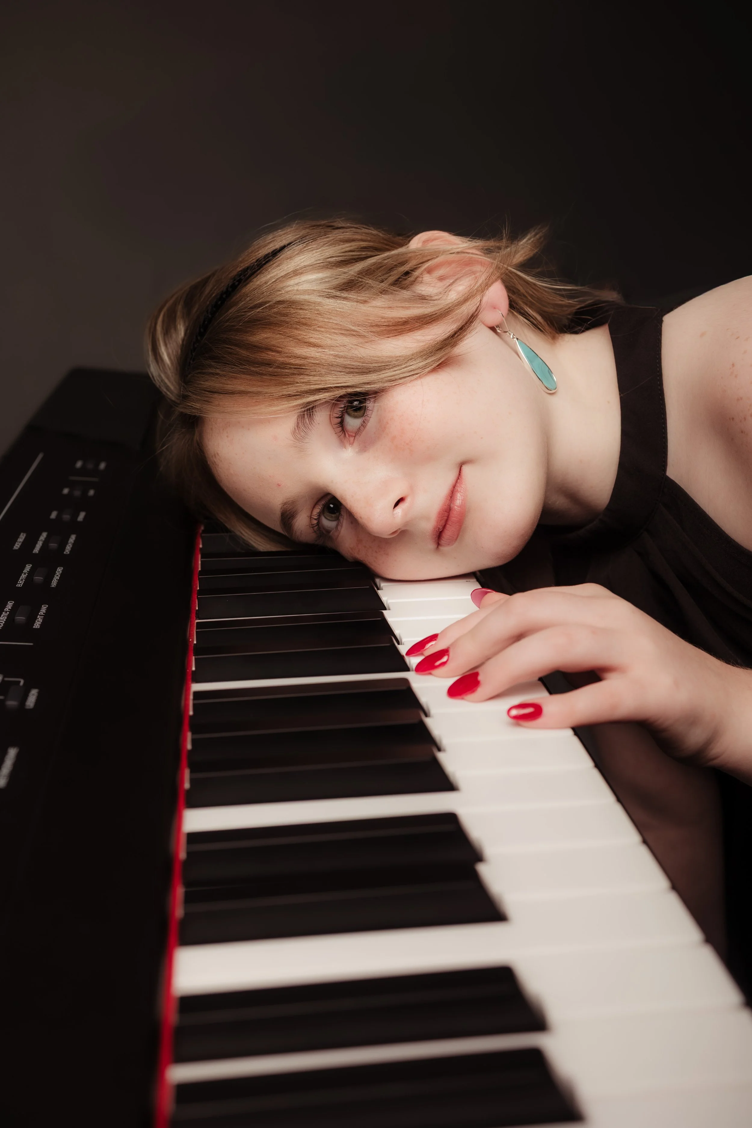 Young woman with light brown hair and teal earrings resting her head on a keyboard piano, looking at the camera with a gentle expression.