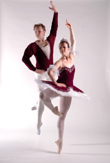 Two ballet dancers, a man and a woman, perform a dance pose together against a plain background. The man stands on one leg, supporting the woman, who is balancing on one leg with her other leg extended upward.