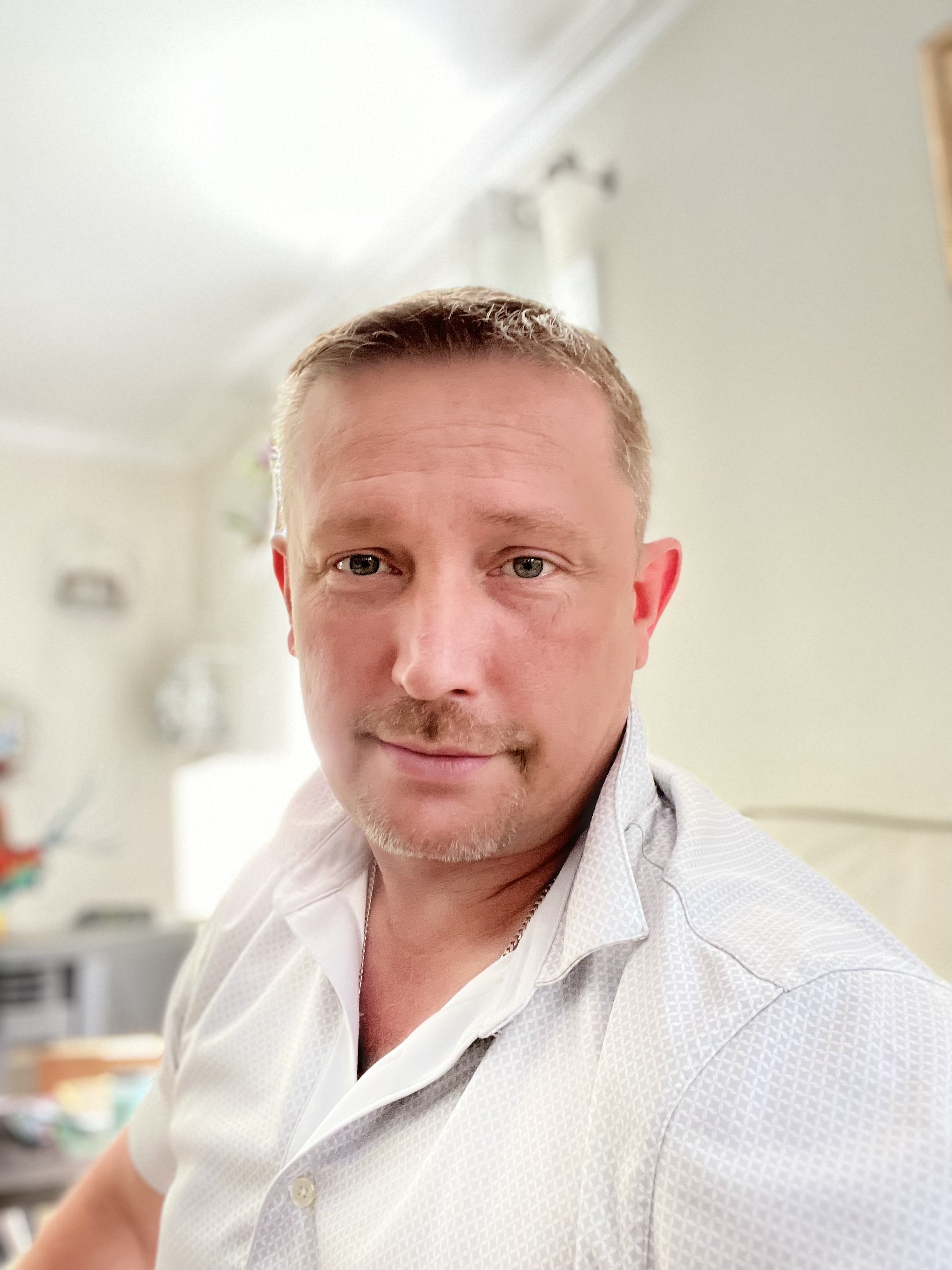 A man with short blond hair, blue eyes, and light facial hair, wearing a white collared shirt, looking into the camera.