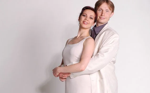 A couple standing close together, smiling, with the woman in a white sleeveless dress and the man in a white jacket and dark shirt against a plain light background.