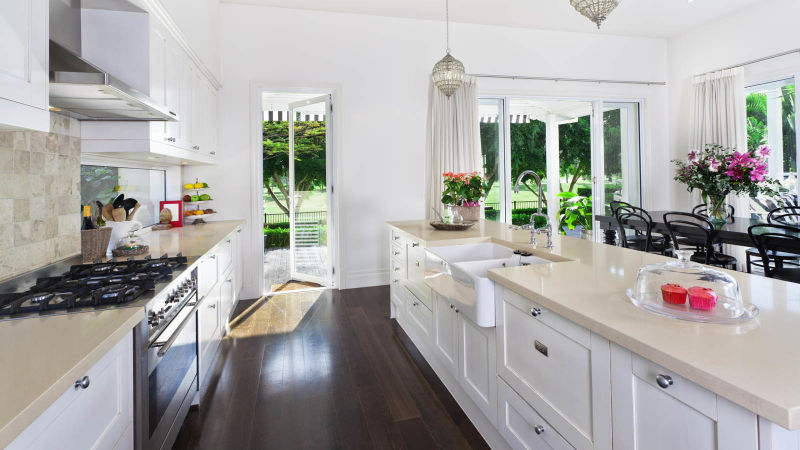 Bright kitchen with white cabinets, dark wooden floors, and a kitchen island with a sink and flowers. Large windows and a glass door lead to a backyard.