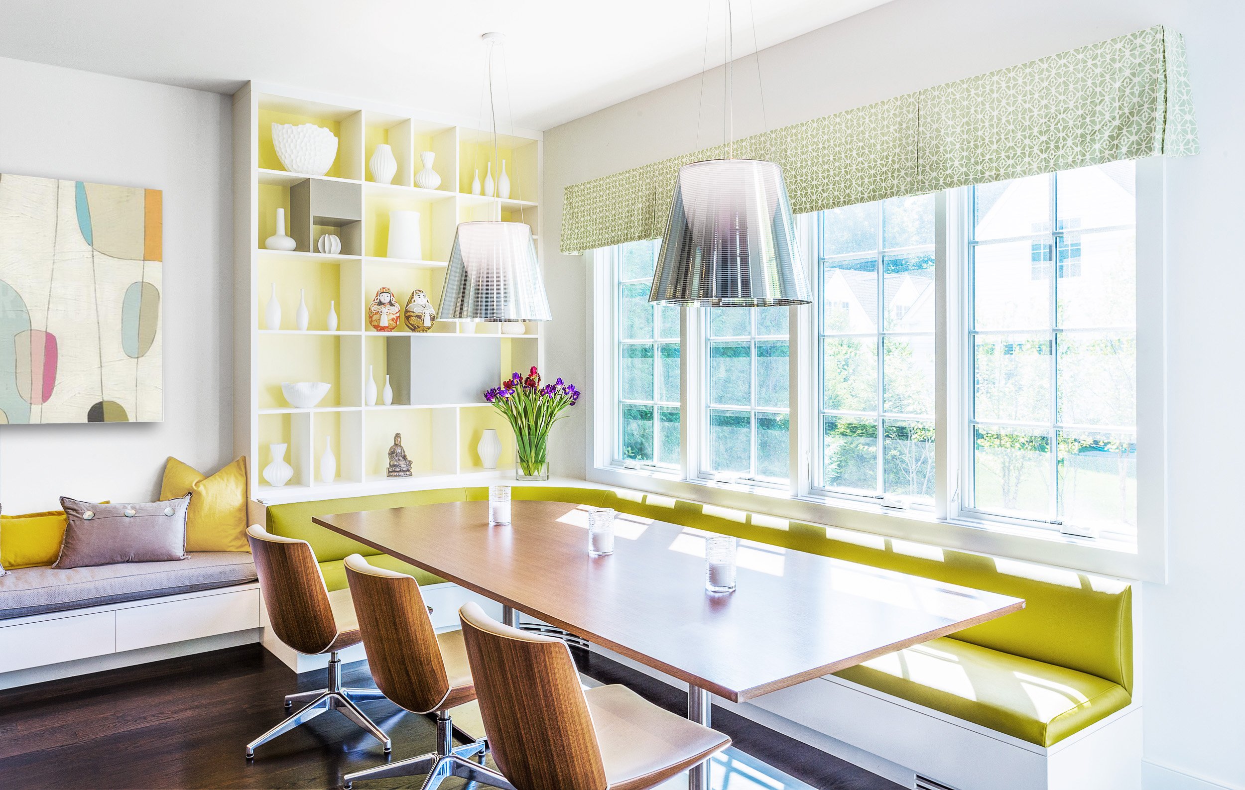 Wide view of a breakfast area with a built-in banquette, lime green cushions, a round-corner dining table, and chrome pendants by the windows in Kensett residence, Darien CT. Designed by Luminosus Designs.