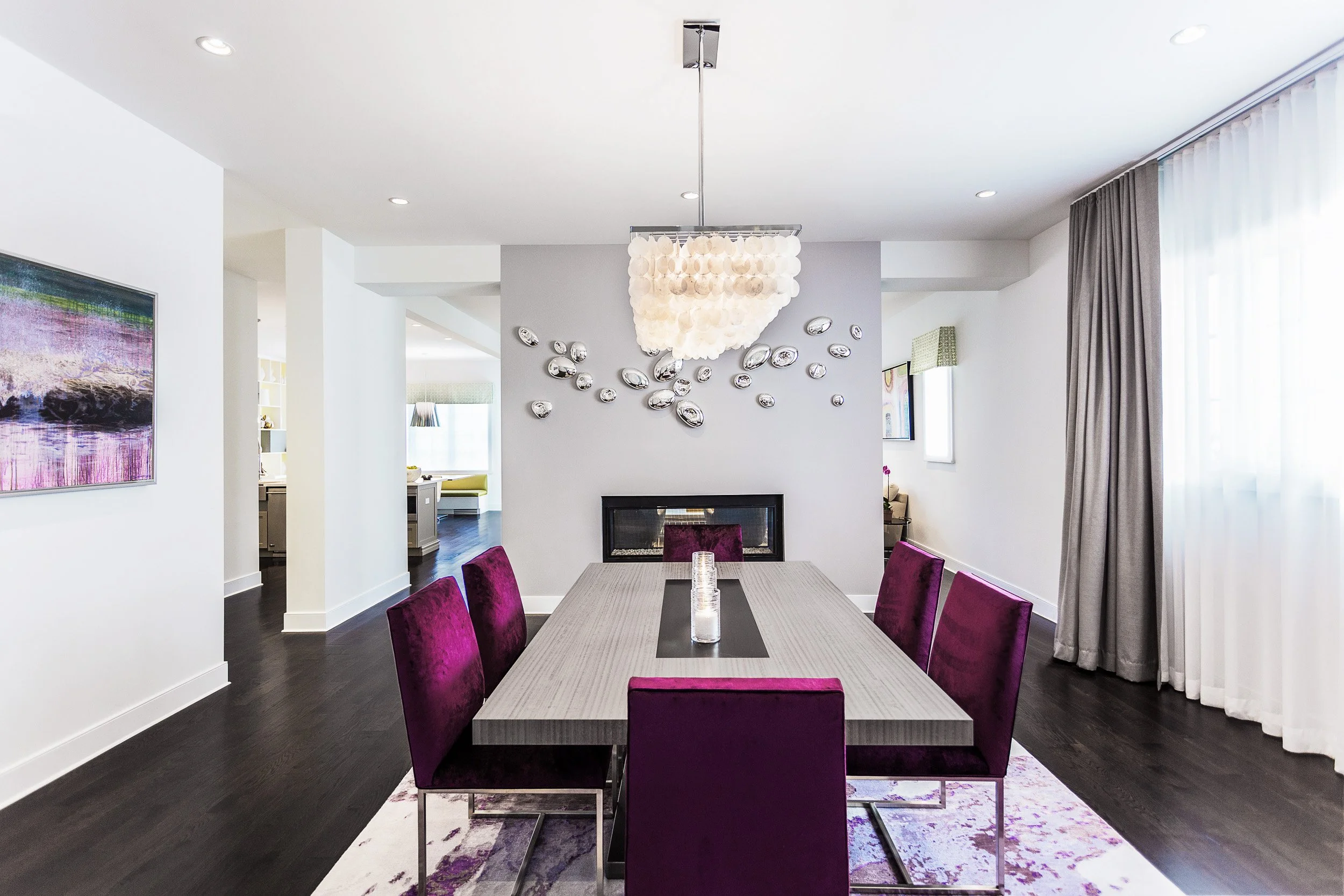 Wide view of a modern dining room with a sleek table, purple velvet chairs, a sculptural ceiling light, and a fireplace backdrop. Designed by Luminosus Designs.