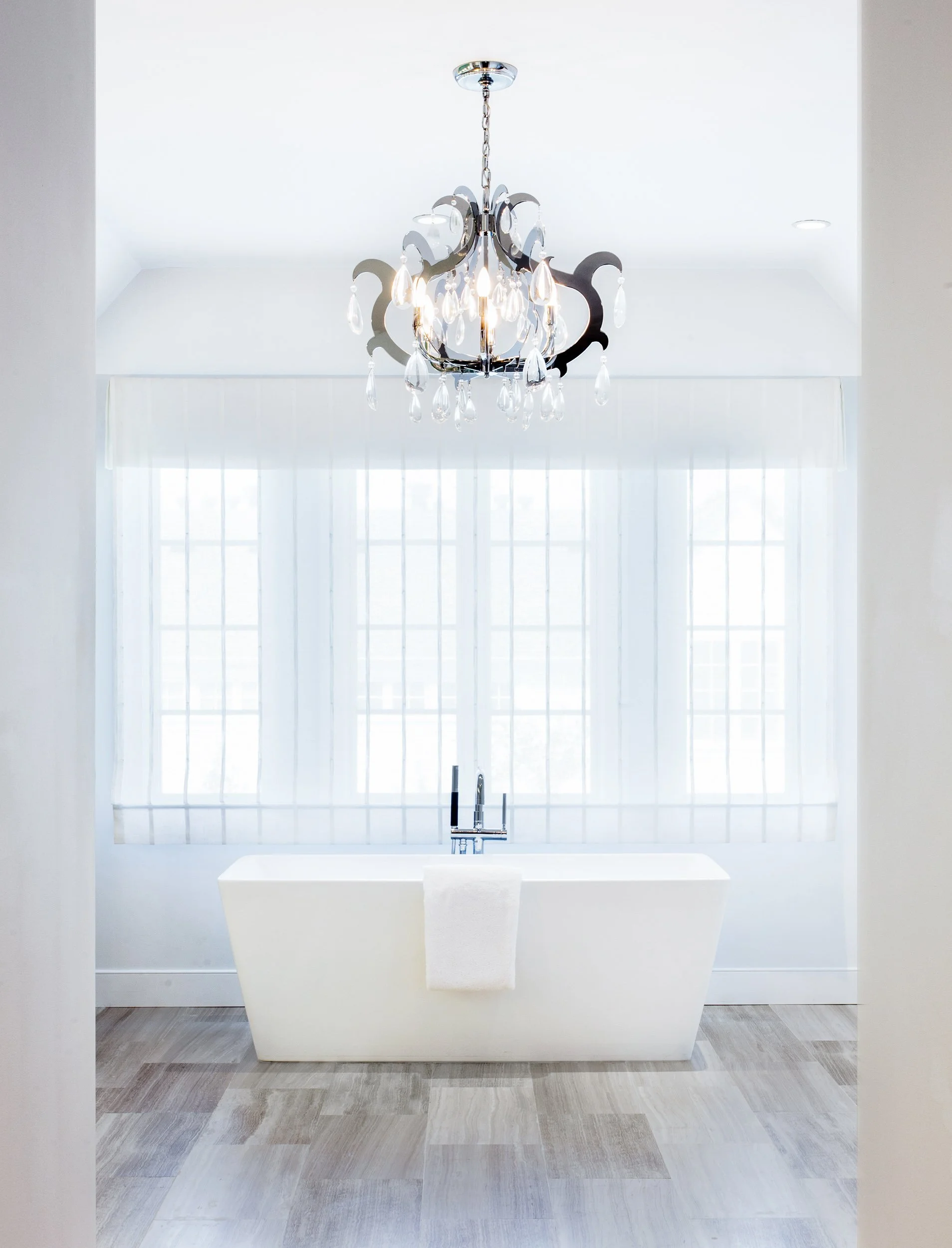 Close-up of a freestanding tub with a floor-mounted filler beneath a crystal chandelier and sheer window treatments.
