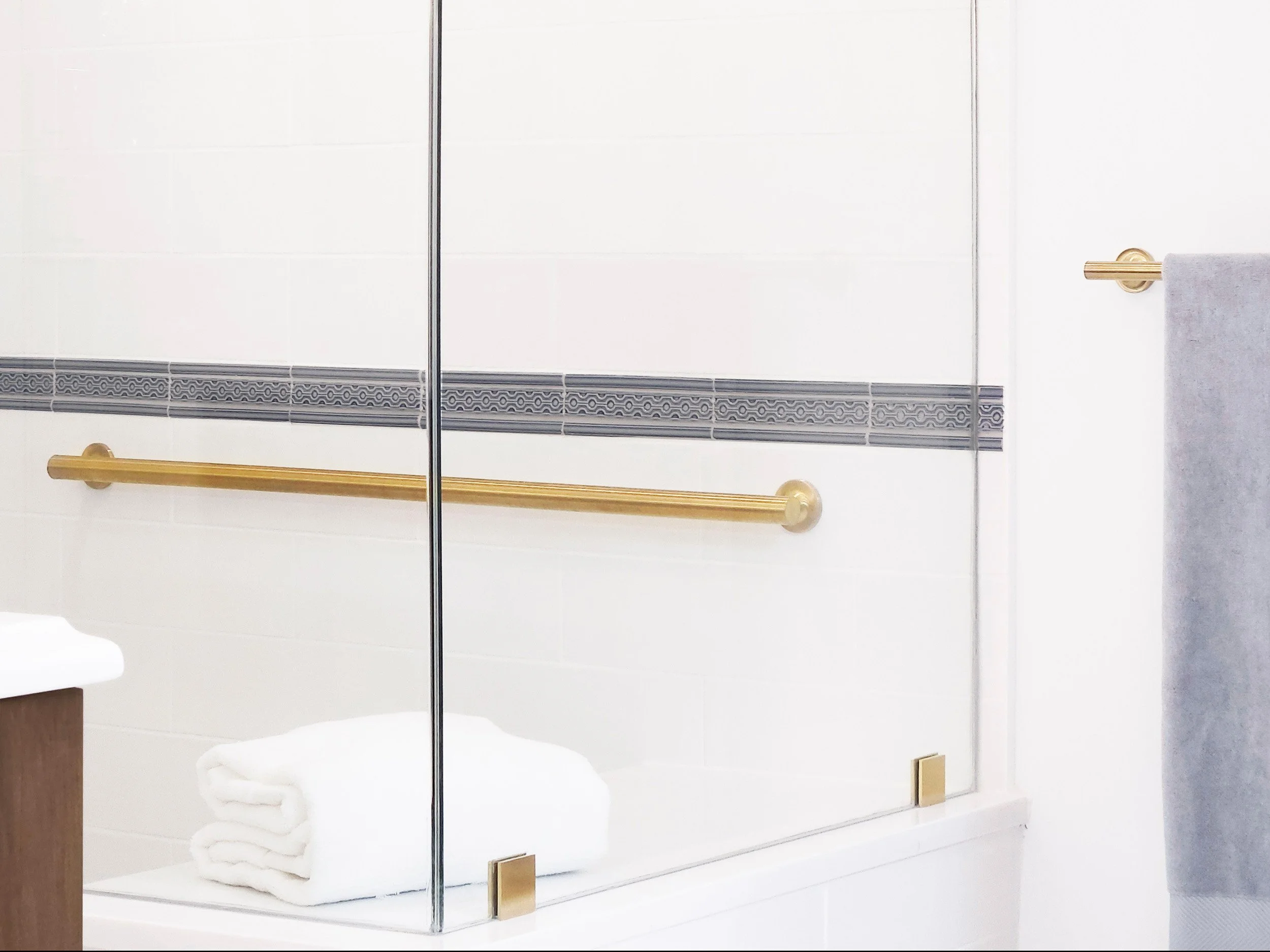 Glass shower corner with folded towels, brass hardware, and grab bar in Princeton, NJ primary bathroom by Luminosus Designs.