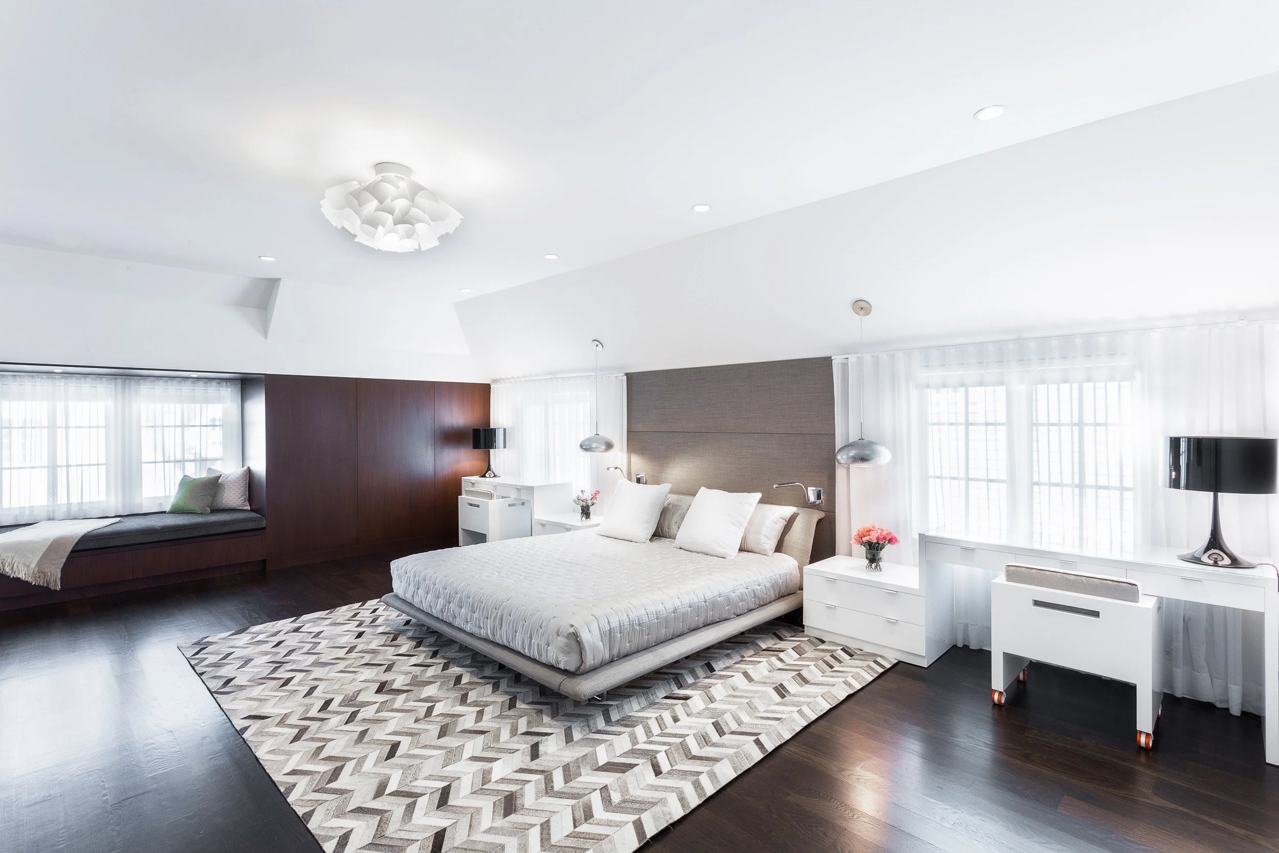 Wide view of a primary bedroom with a platform bed, neutral bedding, chevron area rug, and airy window treatments in Kensett House in Darien, CT. Designed by Luminosus Designs.