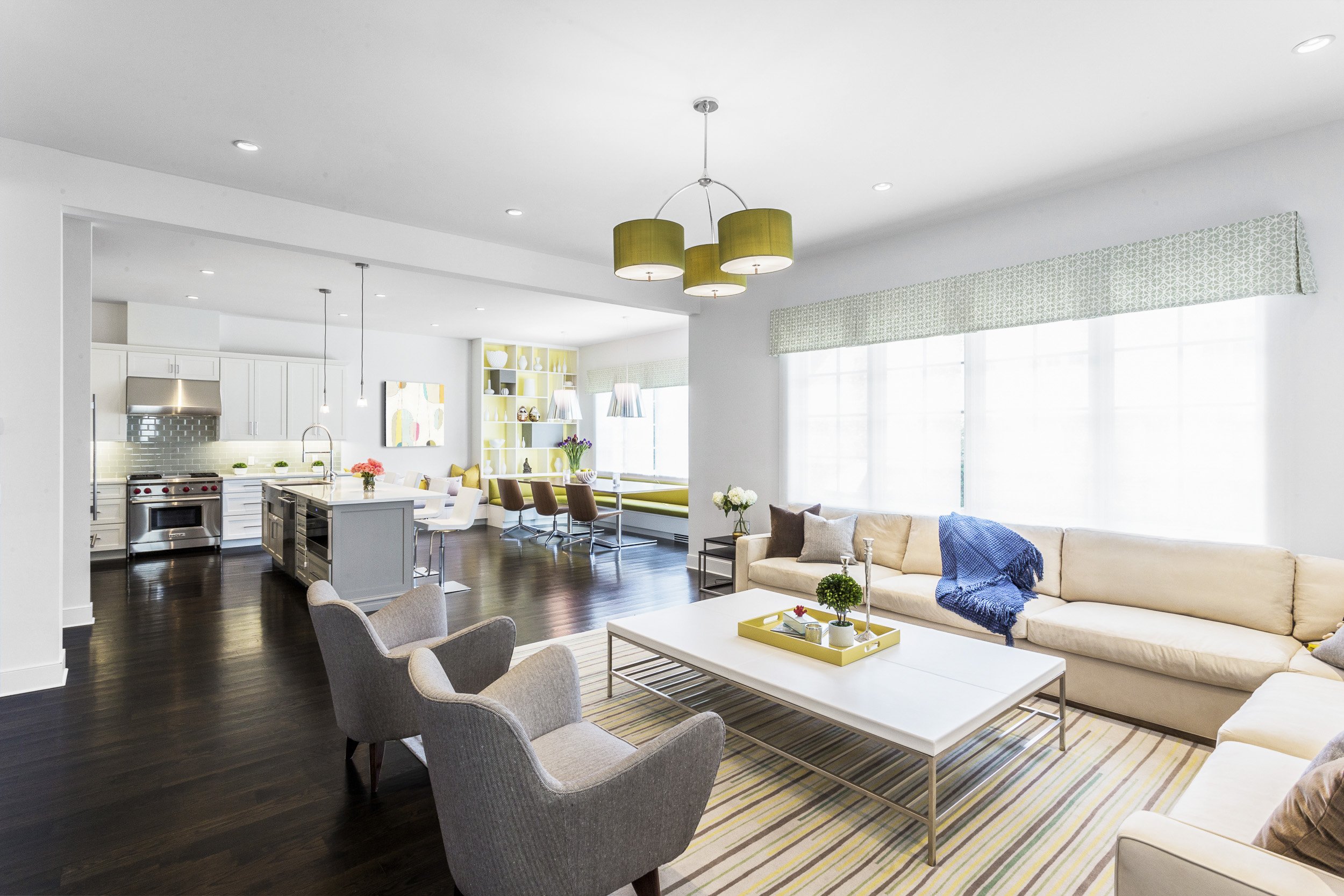 Wide view of an open-concept family room with a tailored sectional, layered pillows, and a statement chandelier with sightlines into the kitchen. Designed by Luminosus Designs.
