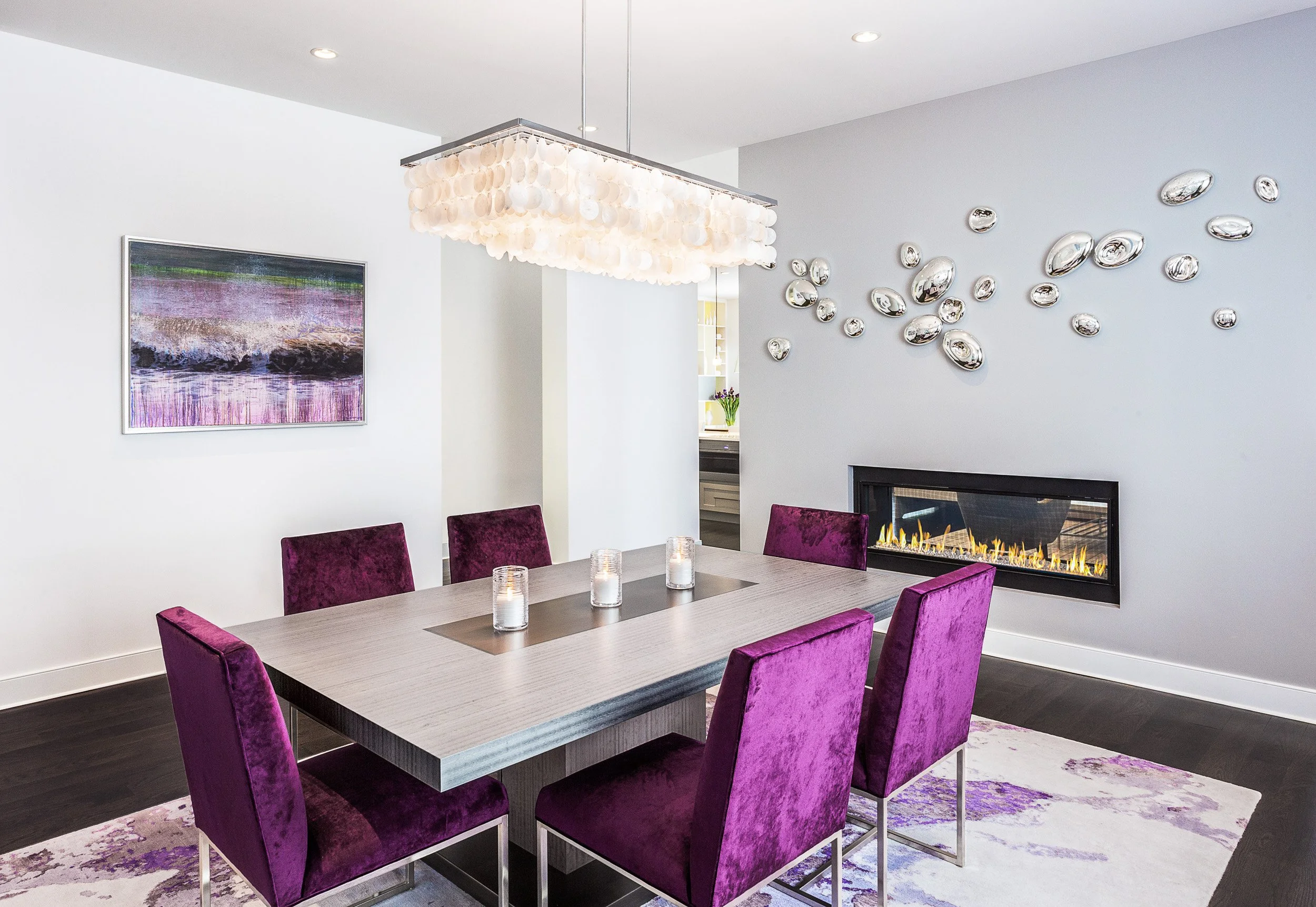 Close-up of the dining table and purple velvet chairs with pendant lighting and fireplace in the background.
