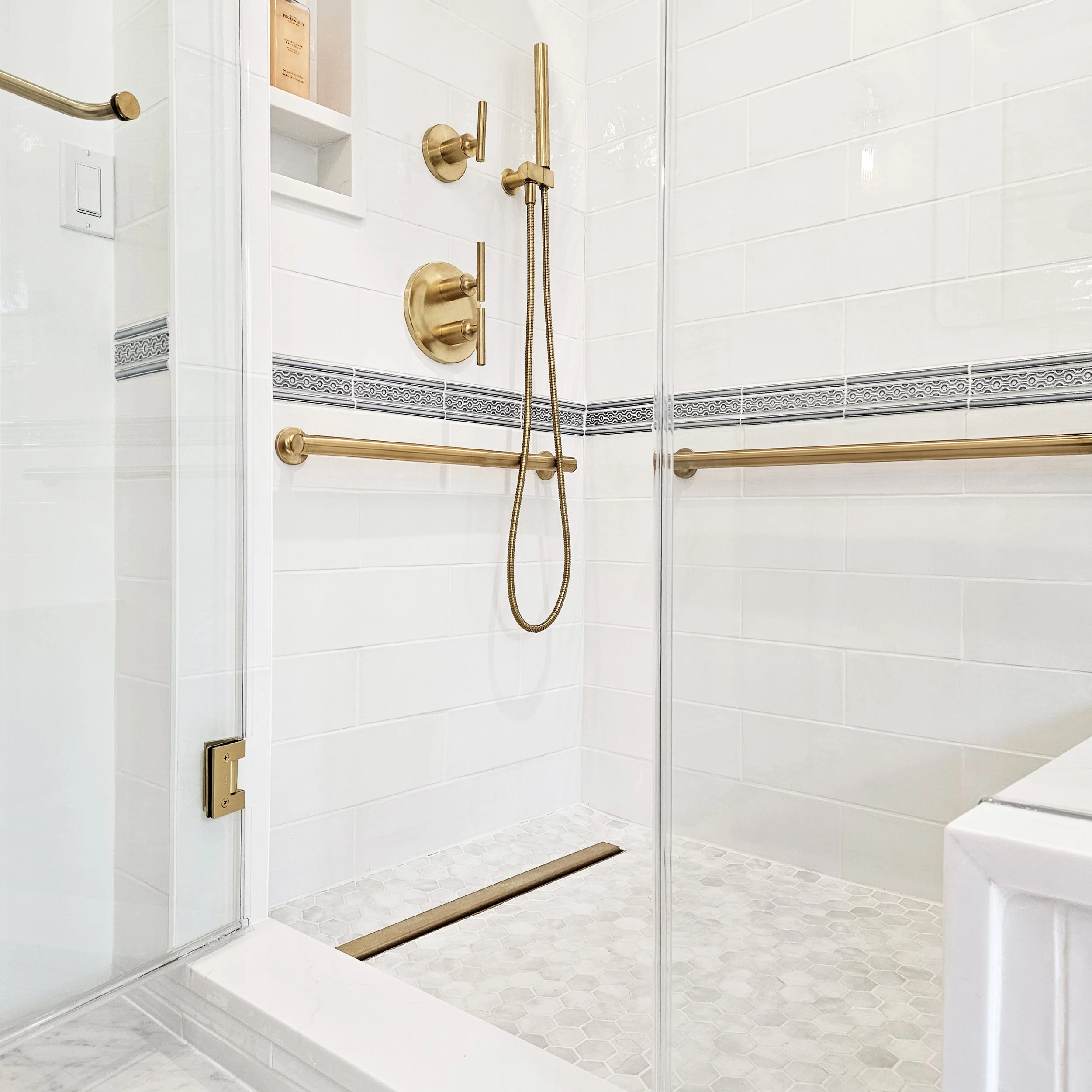 Brass shower fixtures and grab bars against glossy white tile with decorative banding in Carlyle Court in in Princeton, NJ.