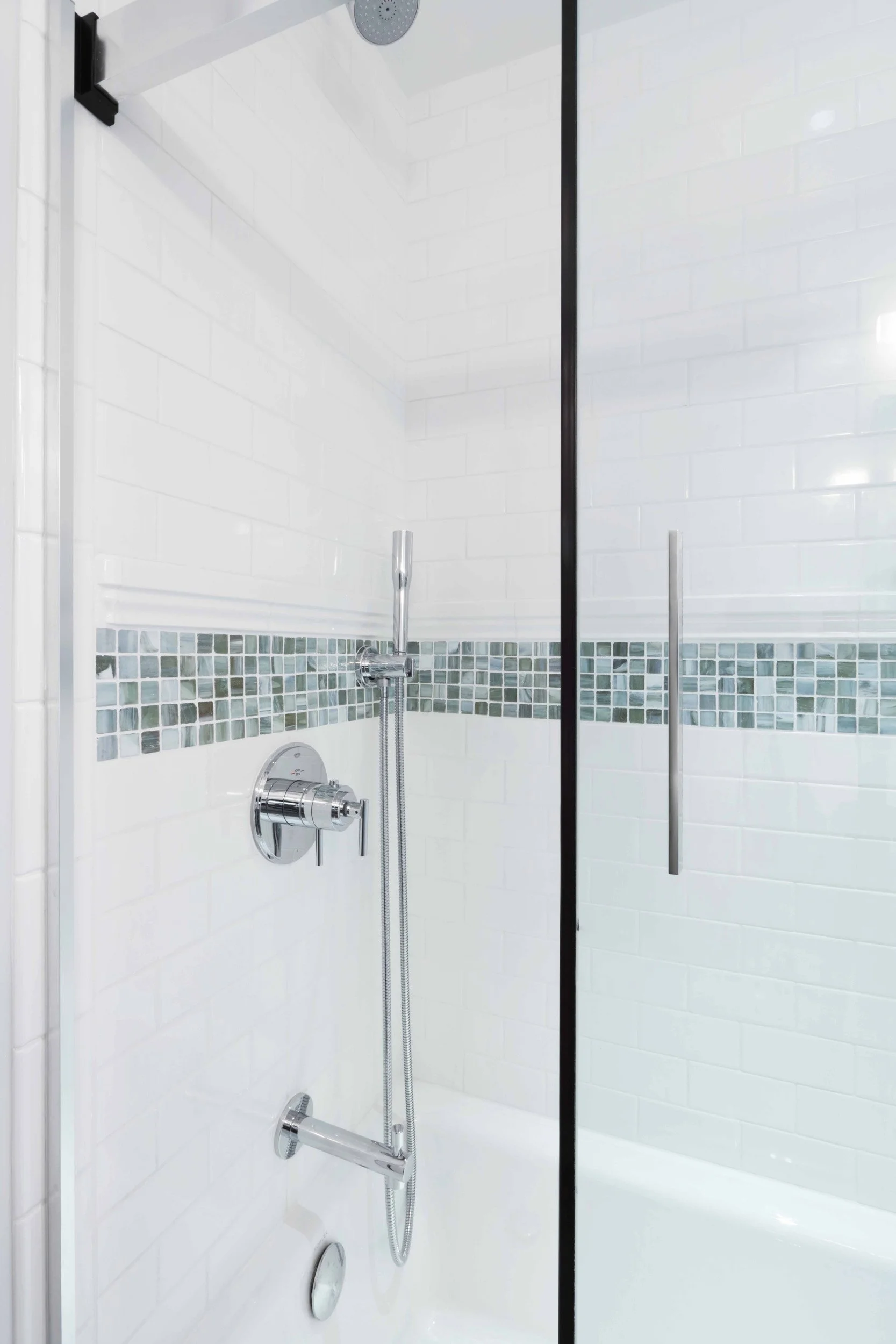 Close-up of a glass-enclosed tub and shower combination featuring white subway tile, mosaic accent band, and chrome fixtures in this family bathroom.