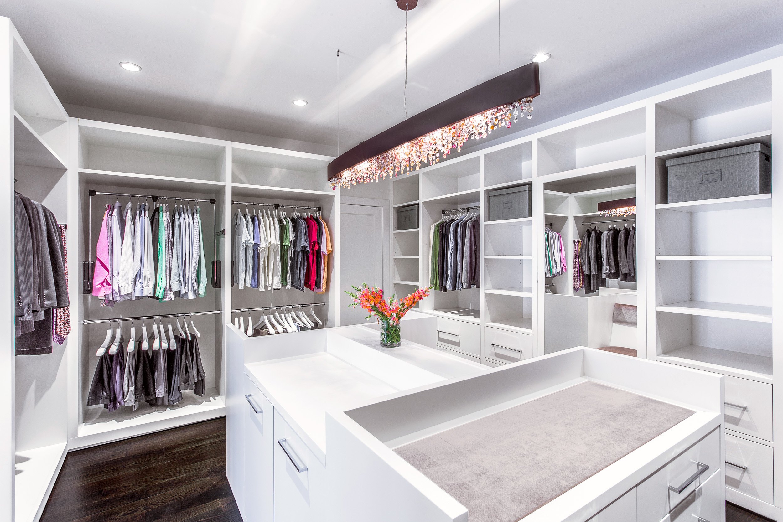 Wide view of a custom walk-in closet with a center island, tailored storage, and a statement crystal chandelier. Designed by Luminosus Designs.