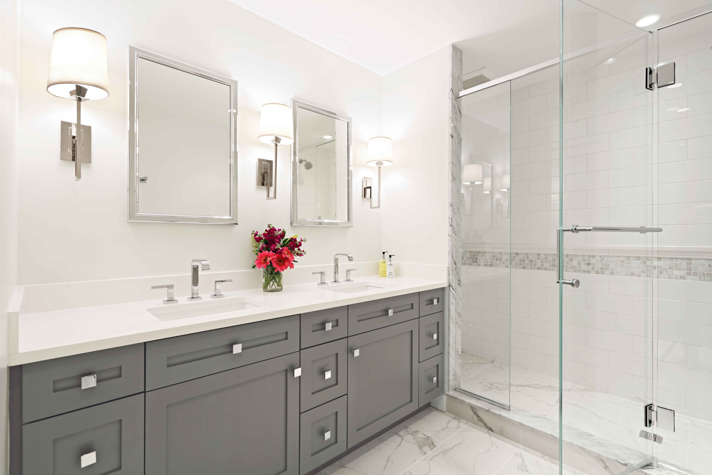 Hero view of a bright primary bathroom featuring a custom gray vanity, quartz countertops, chrome fixtures, and glass-enclosed shower designed by Luminosus Designs.
