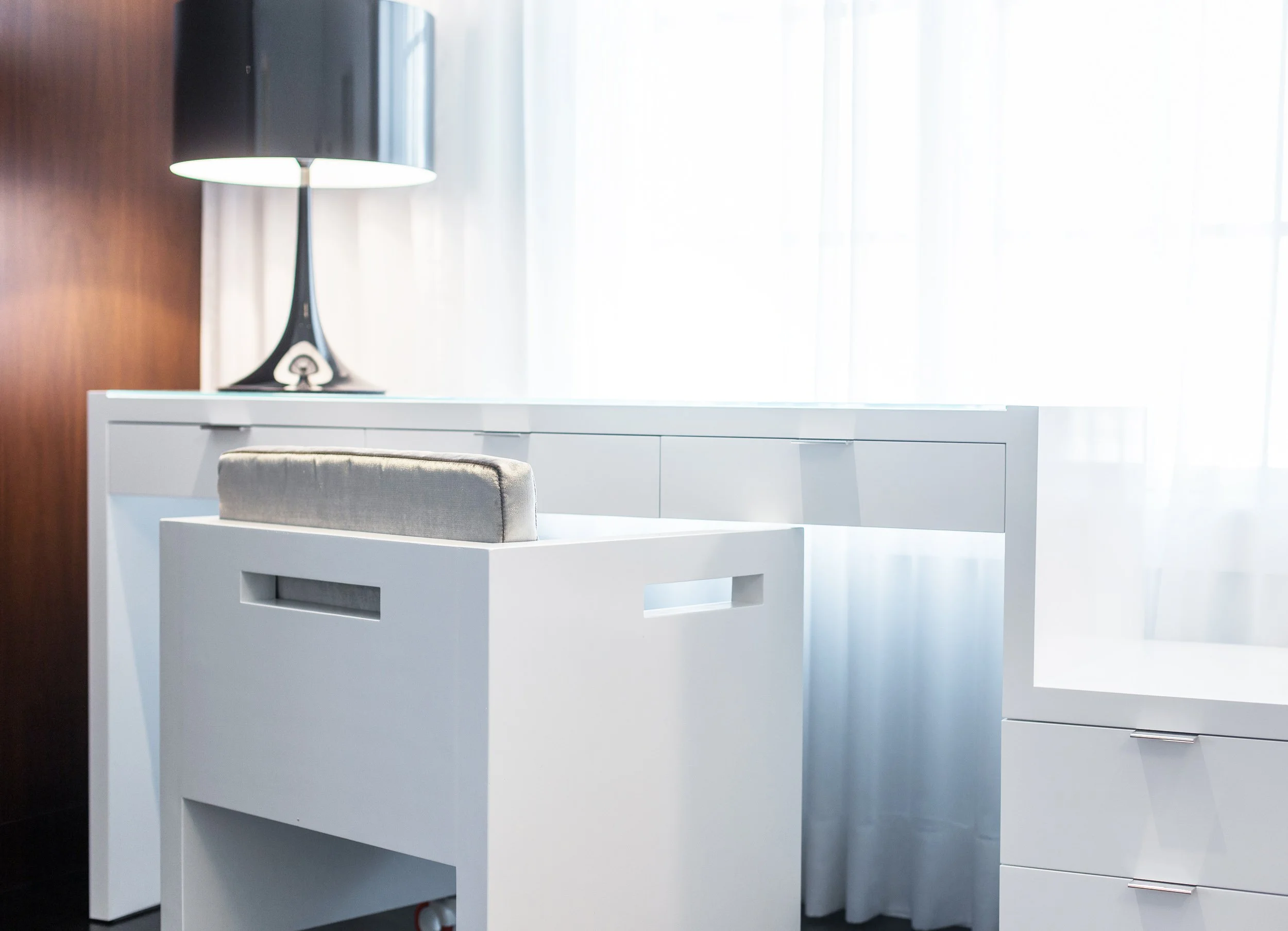 Detail shot of a minimal desk vignette with a white desk, upholstered chair, and black table lamp by the window.
