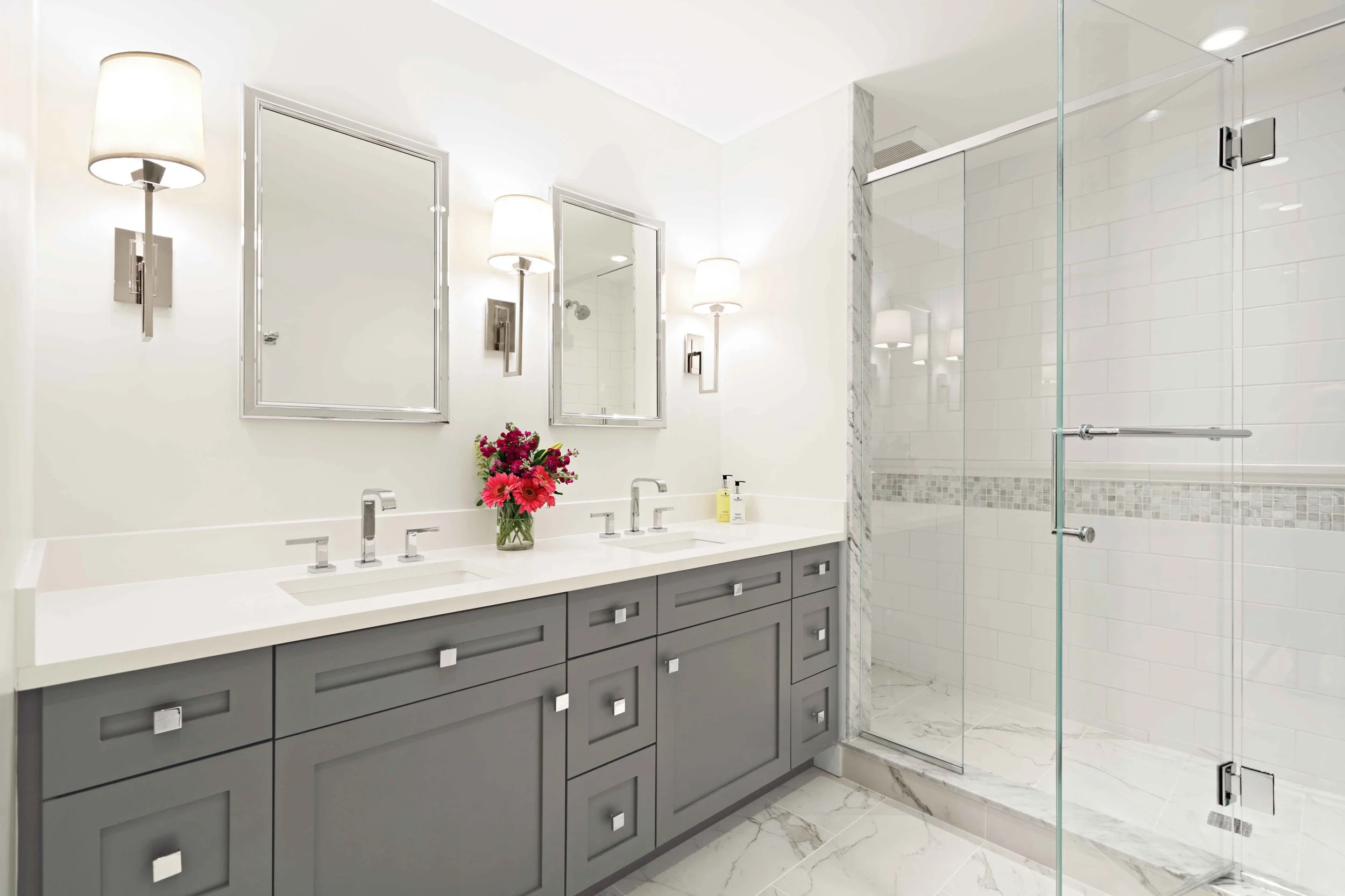 Hero view of a bright primary bathroom featuring a custom gray vanity, quartz countertops, chrome fixtures, and glass-enclosed shower designed by Luminosus Designs.