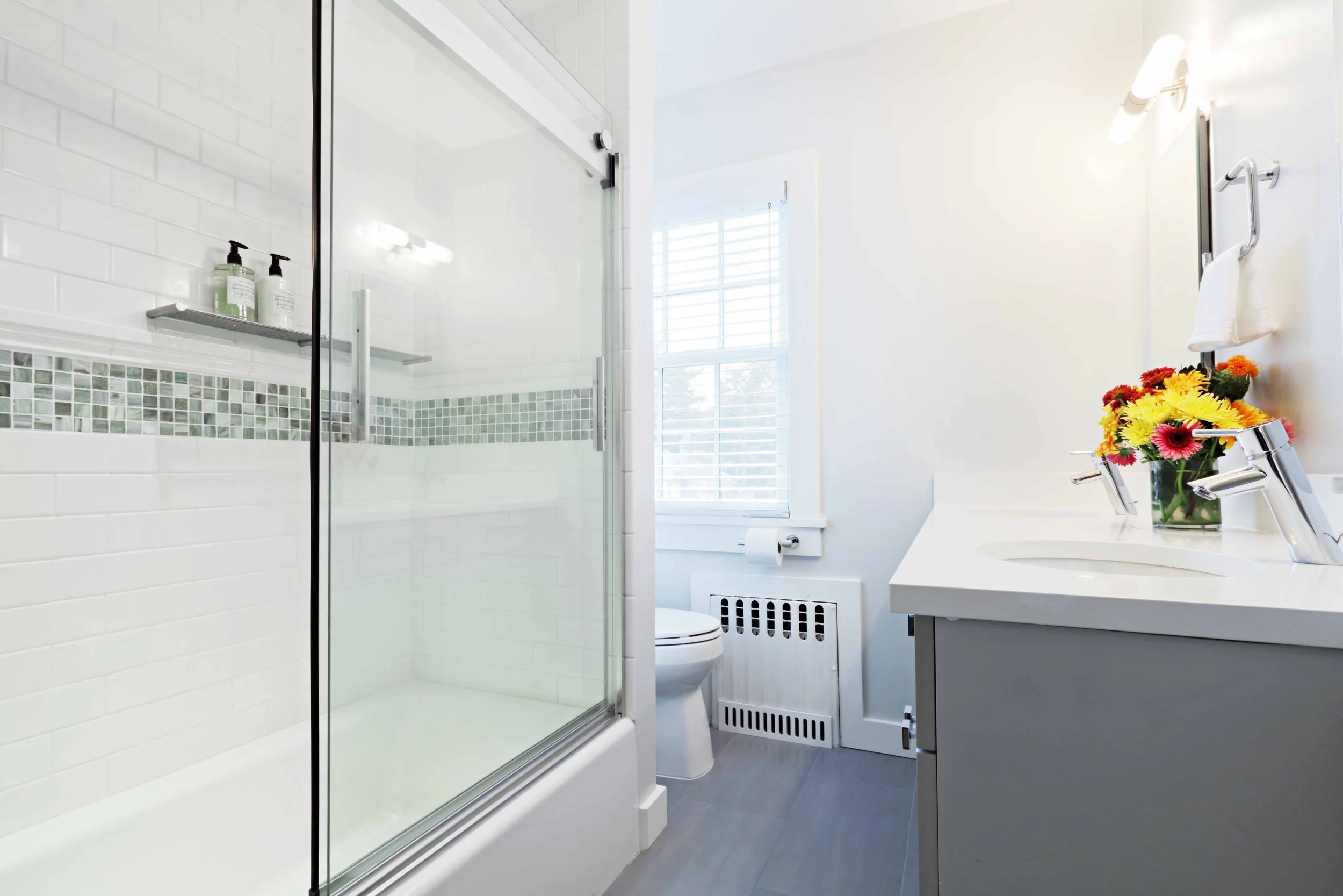 Wide view of a bright family bathroom with glass-enclosed tub shower, white subway tile, mosaic accent band, and custom vanity in Scarsdale, NY.