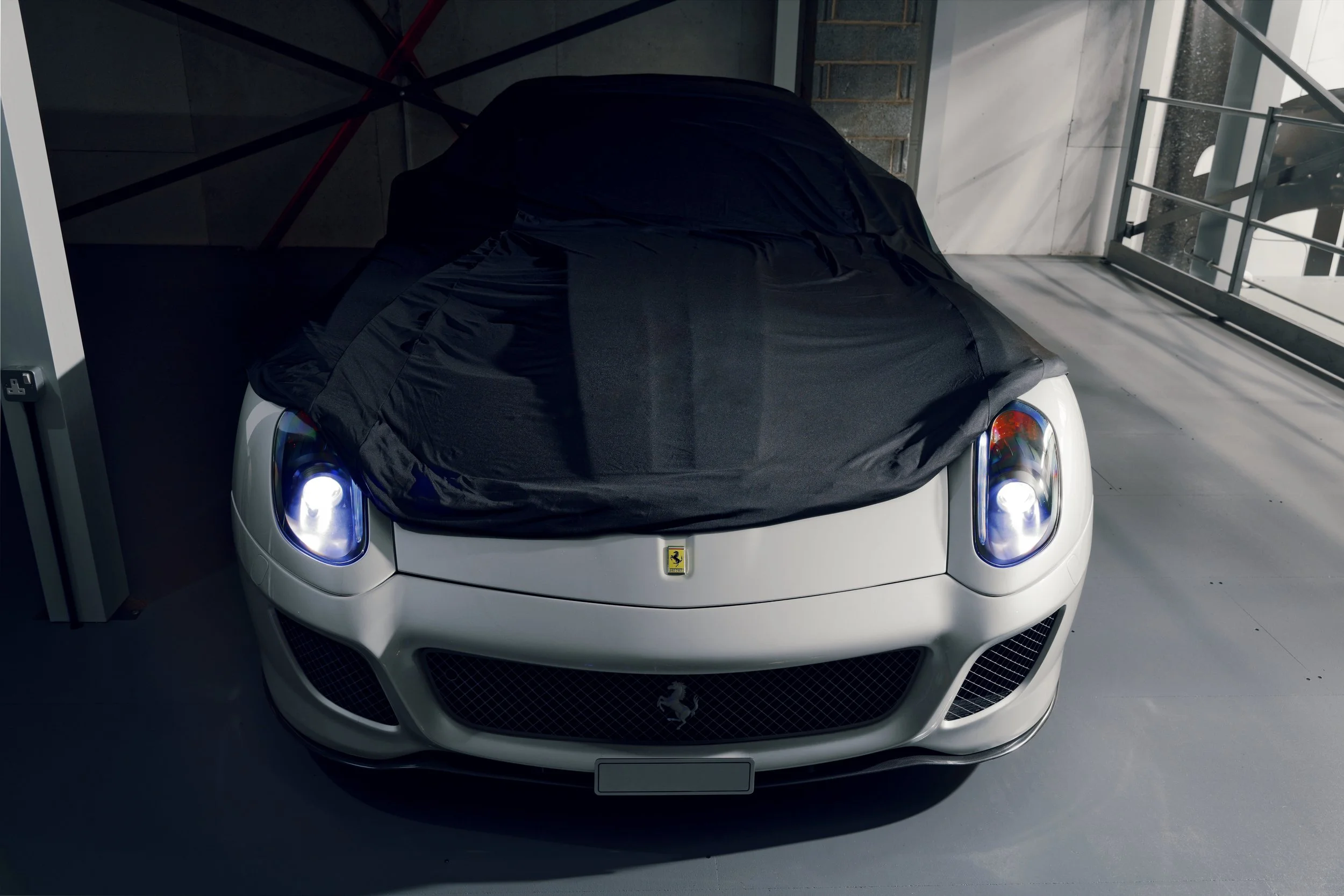 A white Ferrari sports car partially covered with black fabric in a modern indoor parking garage with concrete floors, glass walls, and metal railings.