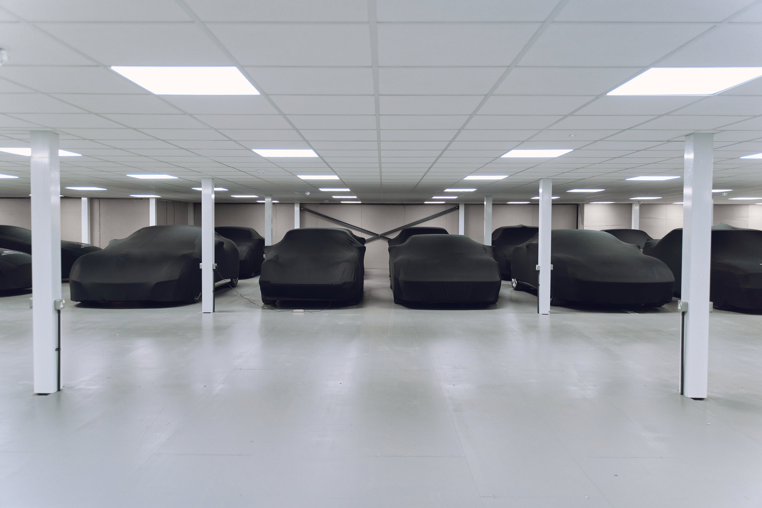 Underground parking garage with multiple cars covered by black car covers, illuminated by ceiling lights, with white support columns.