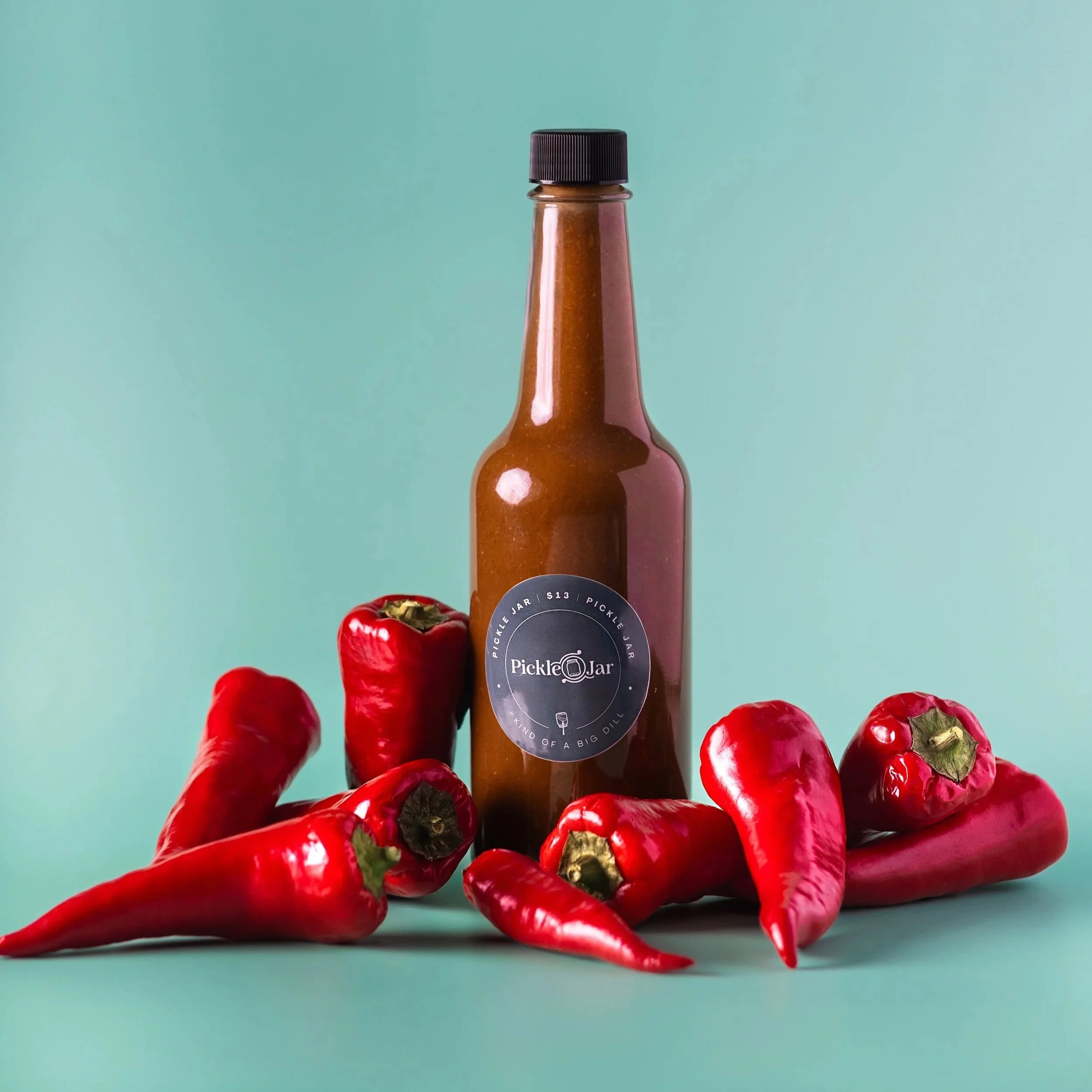 A glass hot sauce bottle labeled 'Pickle Jar' surrounded by red chili peppers on a teal background.
