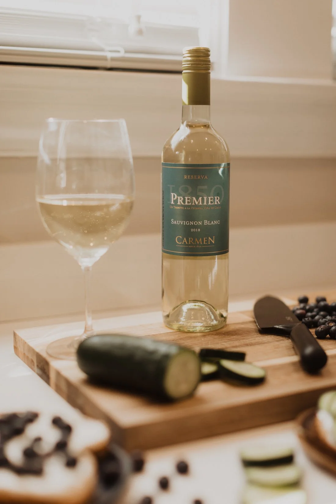 A bottle of Sauvignon Blanc wine next to a glass filled with white wine, a cucumber, and sliced cucumber on a wooden cutting board, with a knife and grapes in the background.