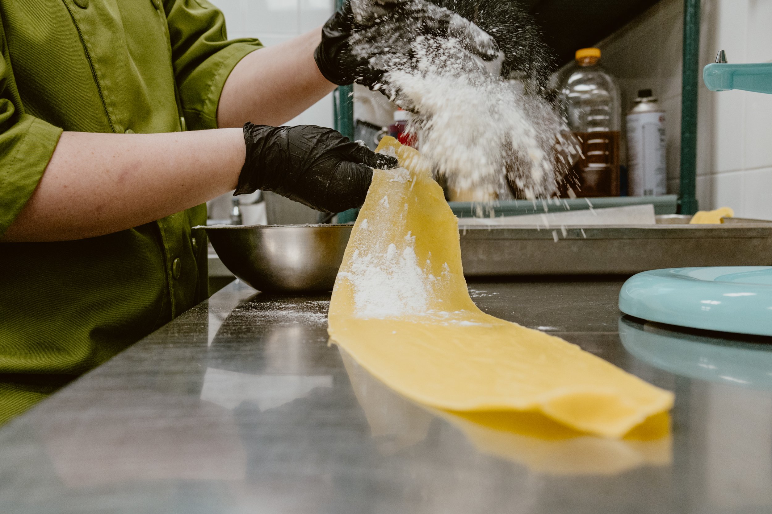 Chef Sarah of Pickle Jar in a green shirt wearing black gloves, preparing fresh pasta dough in Pickle Jar's kitchen, with flour, a mixing bowl, and kitchen items in the background.