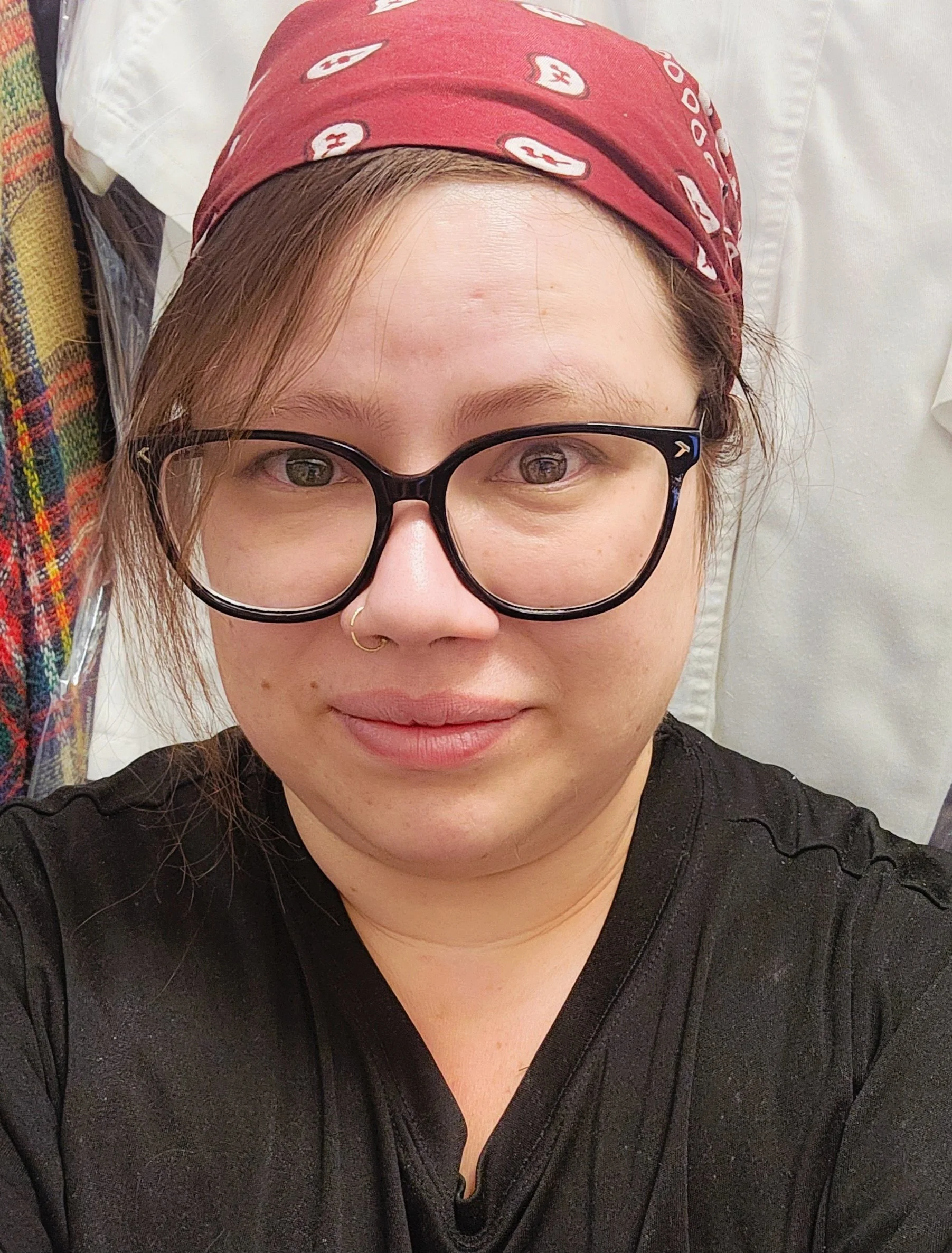 A person with glasses and a red bandana taking a selfie, wearing a black shirt, with a white cloth or curtain in the background.