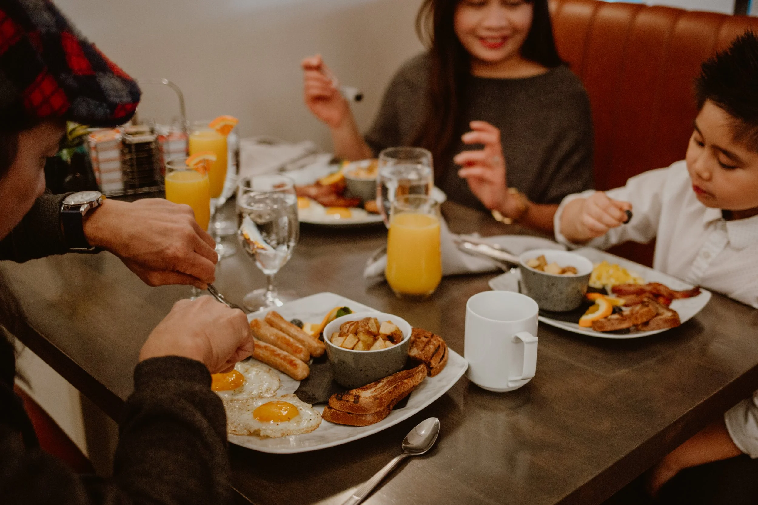 Family having breakfast with eggs, toast, sausages, bacon, orange juice, and water at a table in Pickle Jar restaurant.