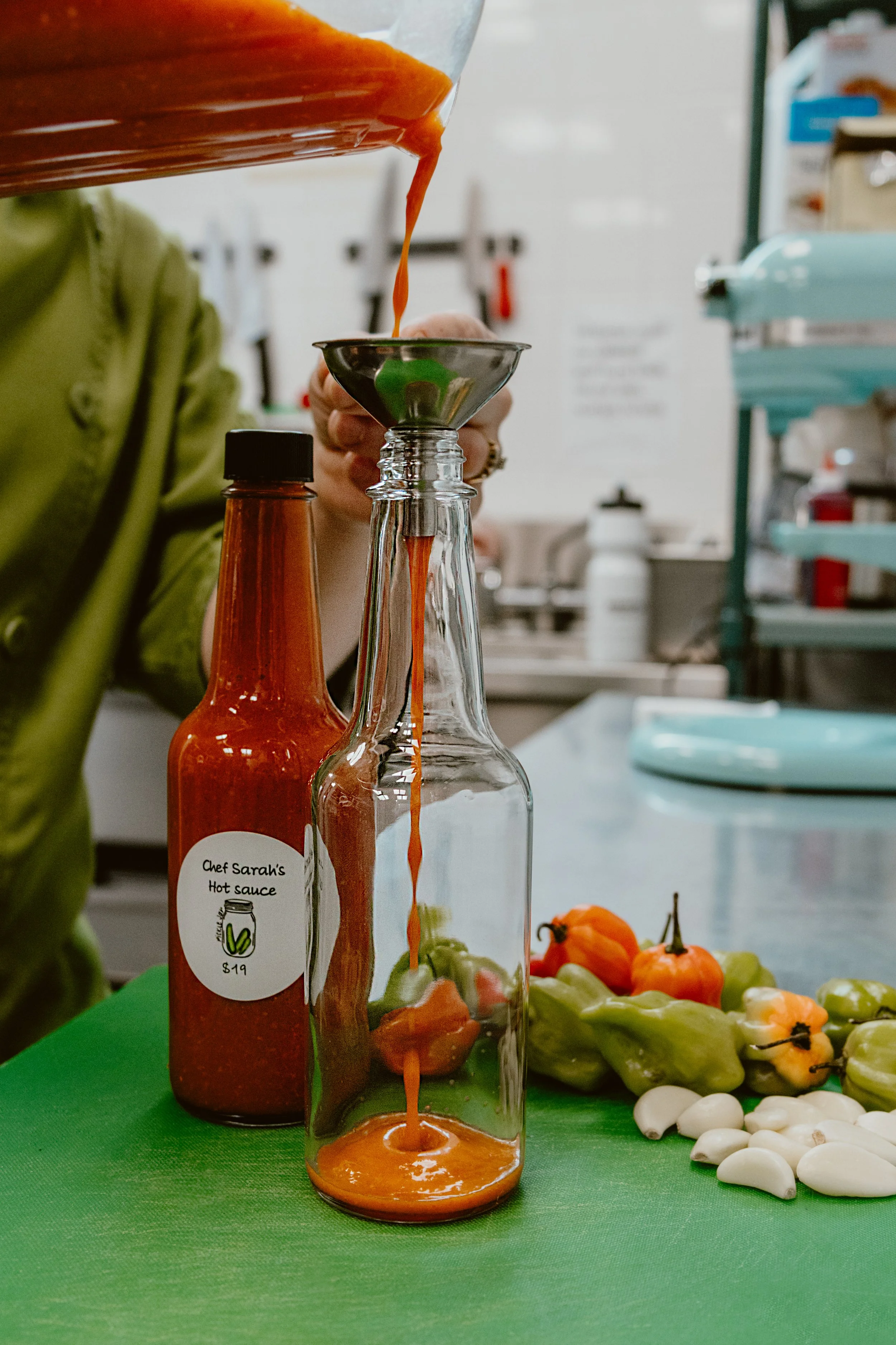 Chef Sarah bottling her freshly created hot sauce with a metal funnel, surrounded by chili peppers and garlic on a green cutting mat in Pickle Jar's kitchen.