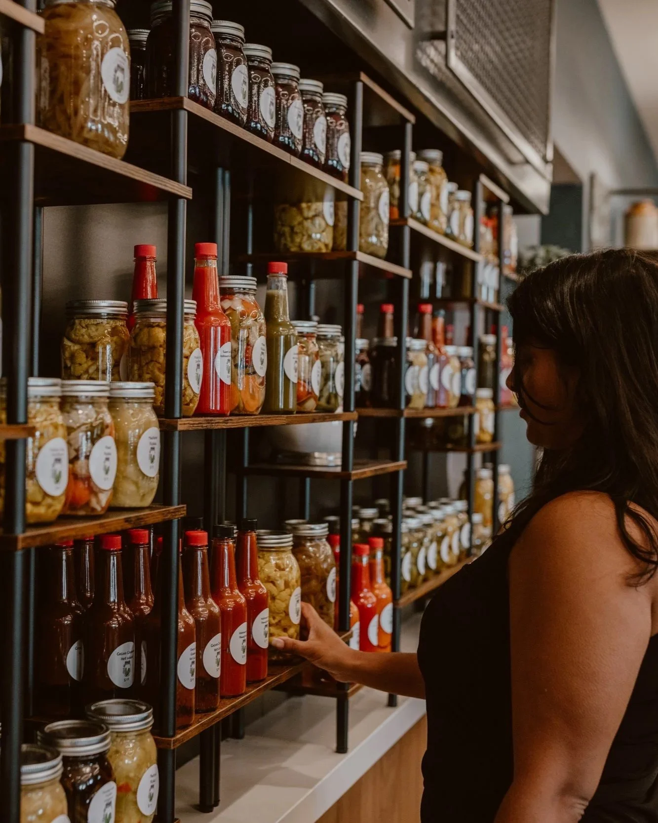 A woman shopping at Pickle Jar's Pantry with shelves filled with jars and bottles of hand made pickled goods and hot sauces for purchase inside the Pickle Jar restaurant.