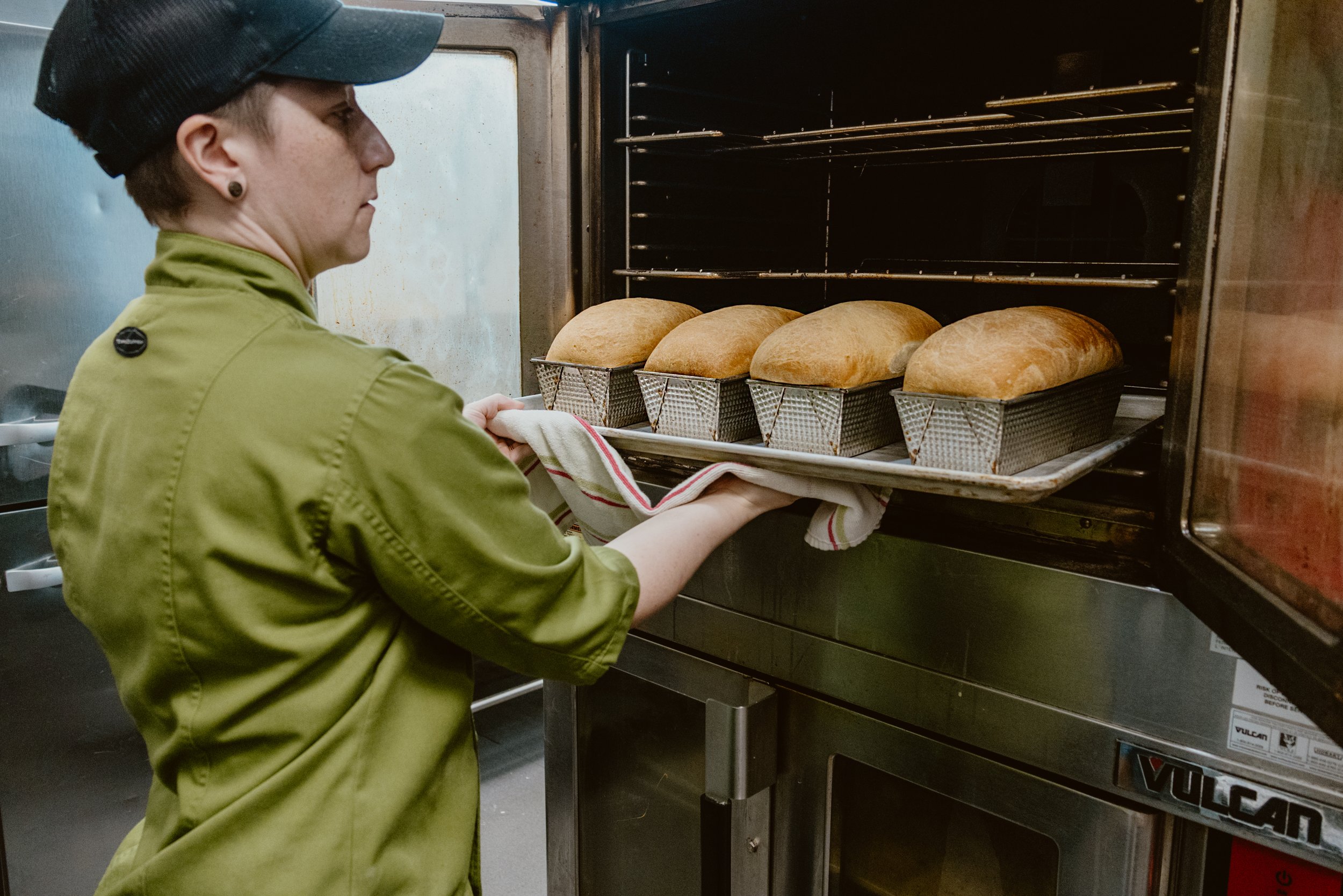 Chef Sarah pulling freshly baked, homemade bread from Pickle Jar's oven. The oven has four loaves of bread cooling in metal baskets on a baking sheet.