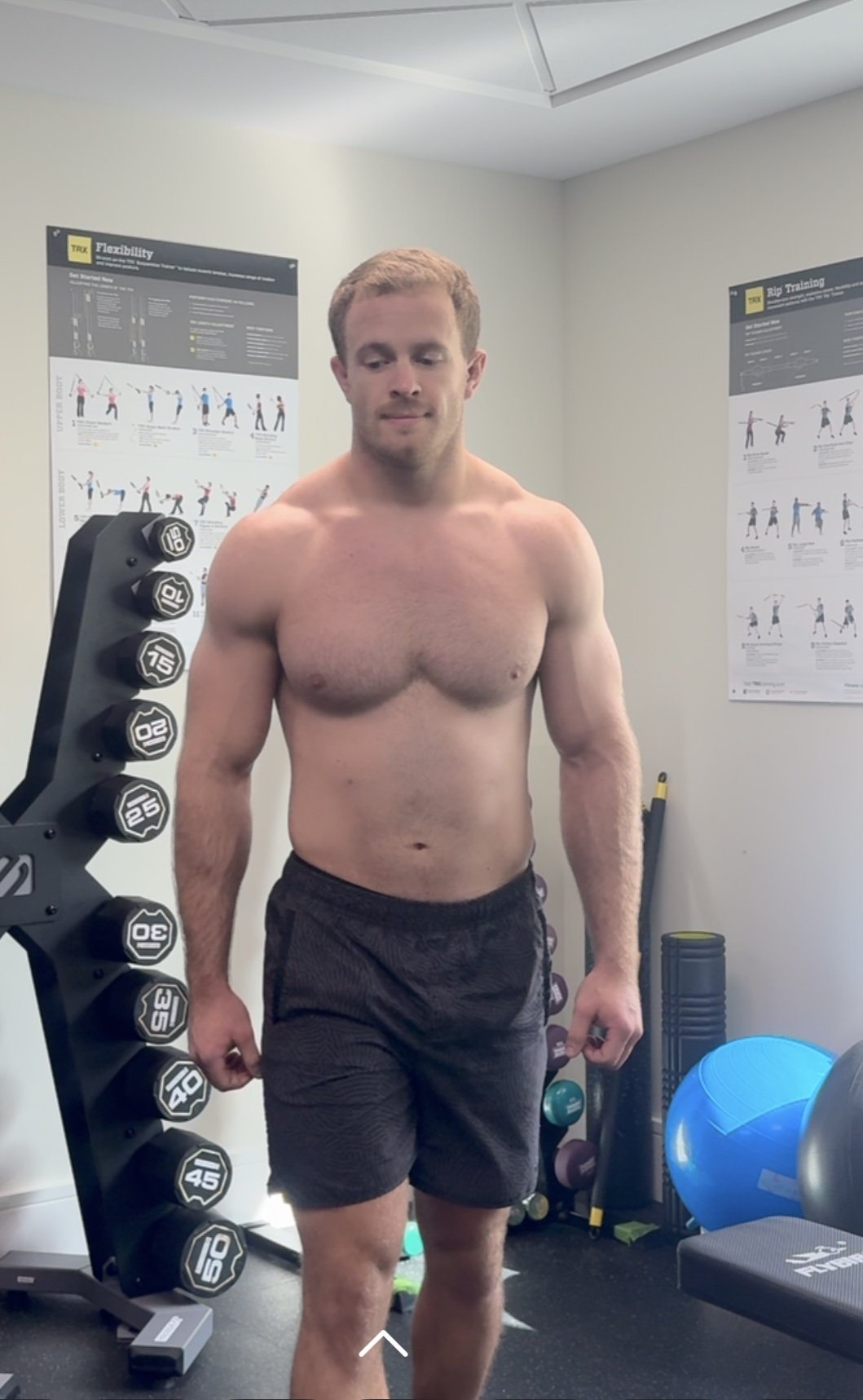 A shirtless man in black shorts standing in a fitness room with dumbbells, exercise posters, and fitness equipment in the background.
