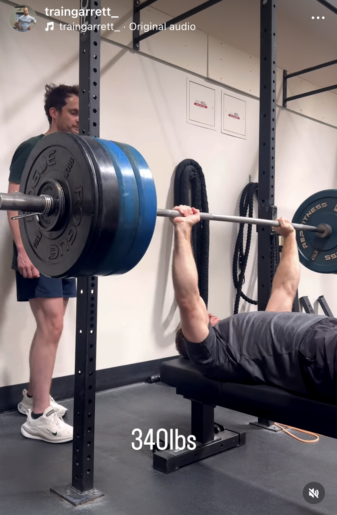A man lying on a bench performing a bench press with a barbell loaded with multiple weight plates, supervised by another man in a gym.
