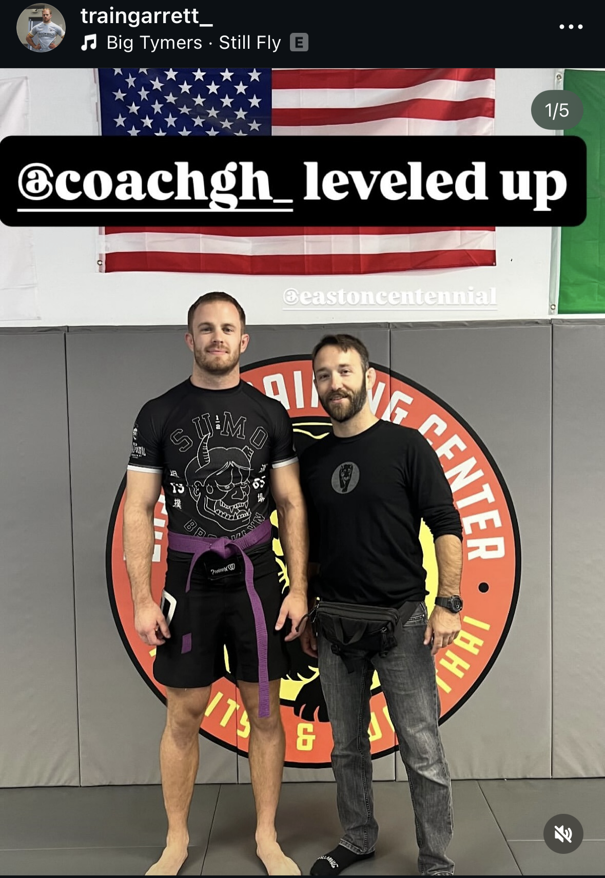 Two men standing in a martial arts gym in front of a logo on the wall. The man on the left is barefoot, wearing a black rash guard with a skull and horn graphic, black shorts, and a purple belt. The man on the right is wearing a black long-sleeve shirt, grey jeans, a watch, and a black waist pouch. The background includes a large American flag and a gray padded wall with a red, yellow, and black circular logo.
