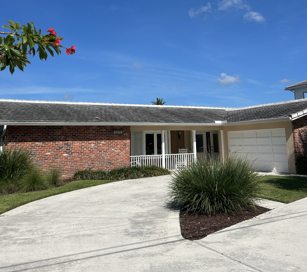 Front view of a single-story brick house with a gray shingled roof, white garage door, and a small porch with a white railing, surrounded by green grass and plants, under a clear blue sky.