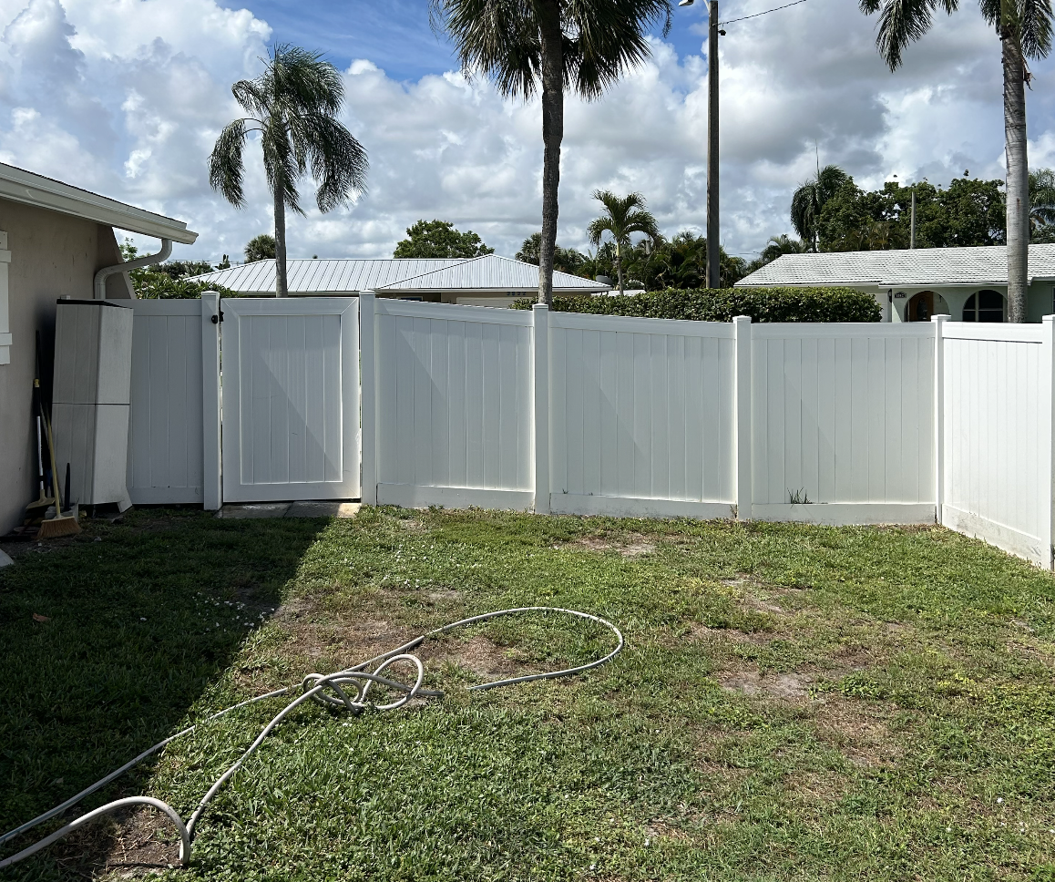 A fenced backyard with a white vinyl fence, patchy grass, a garden hose, and palm trees under a cloudy sky.