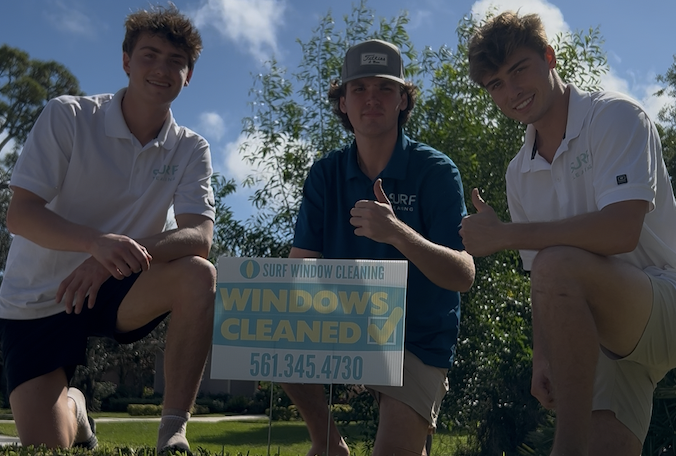 Three young men outdoors next to a sign for a window cleaning business, smiling and giving thumbs up.